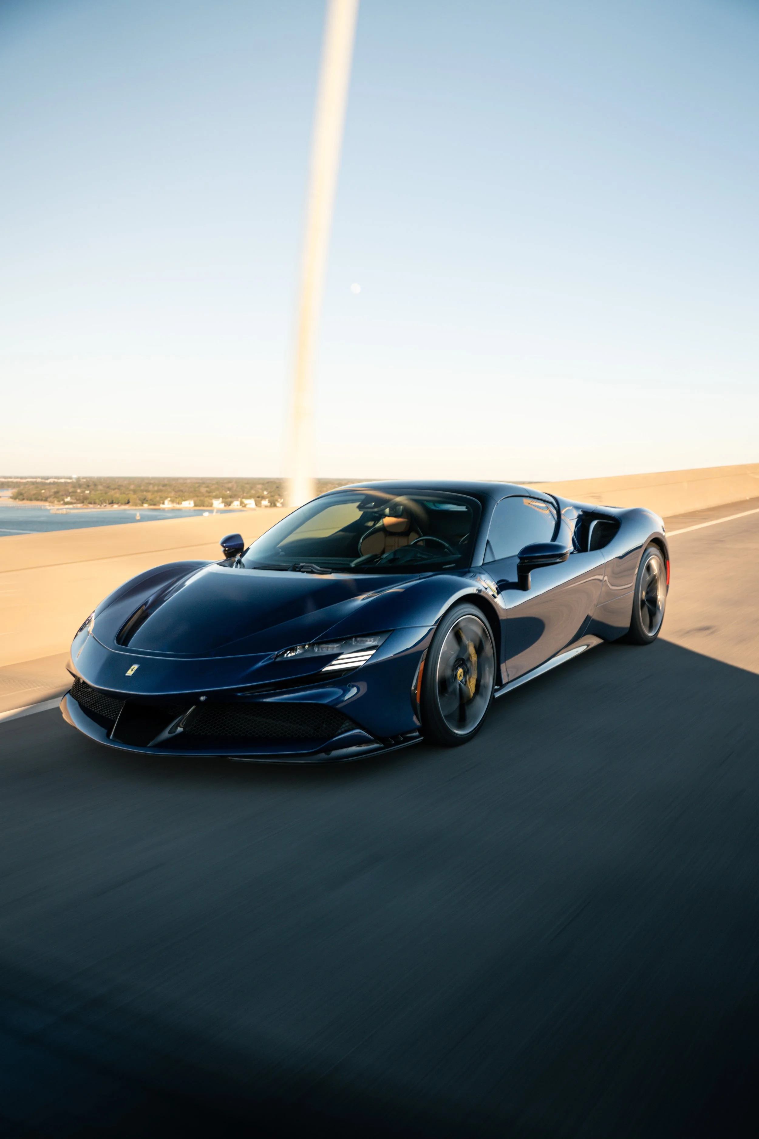 A sleek black sports car driving on a highway with a body of water and a clear blue sky in the background.