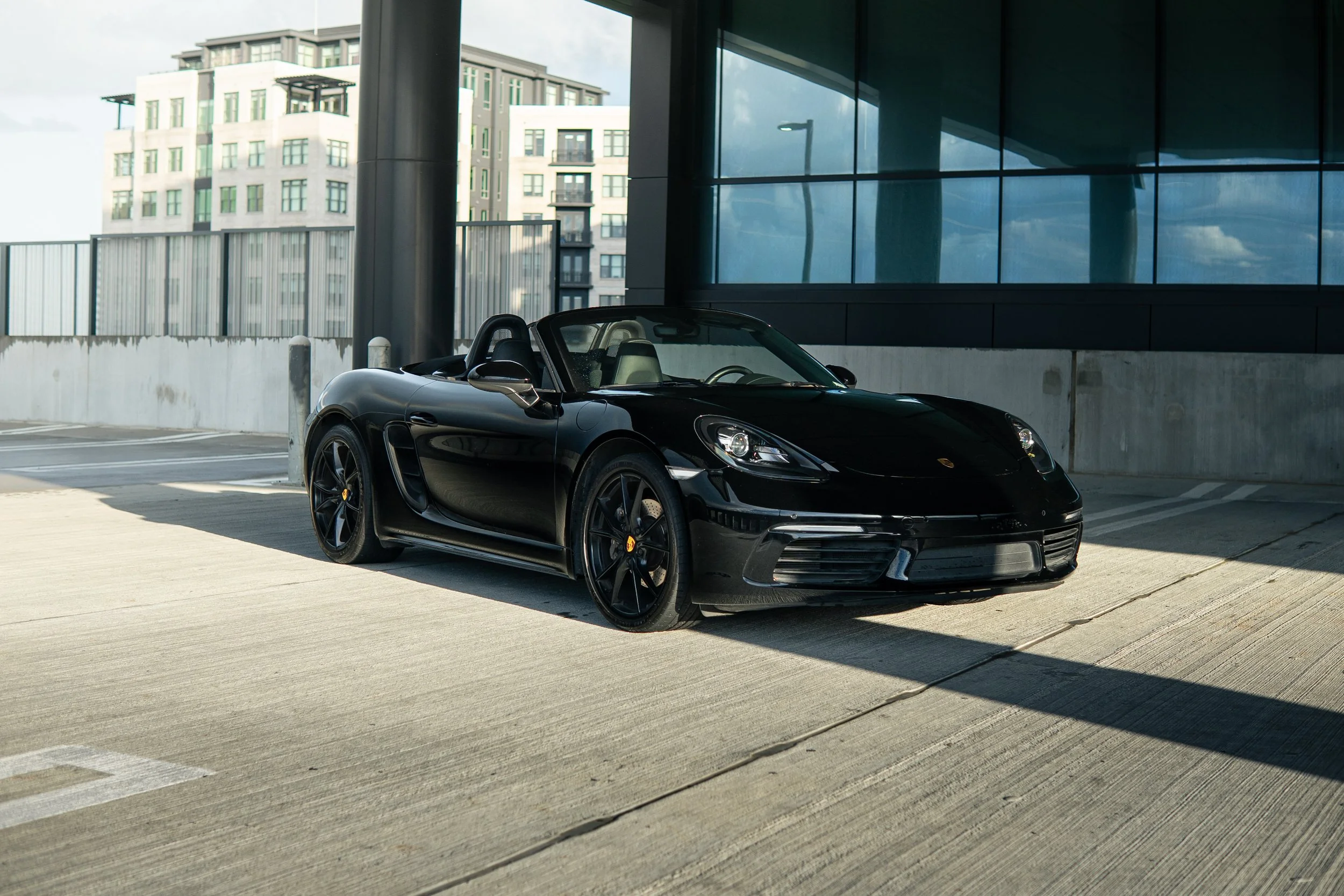 A black Porsche sports car parked in an outdoor parking lot with modern buildings and large glass windows in the background.