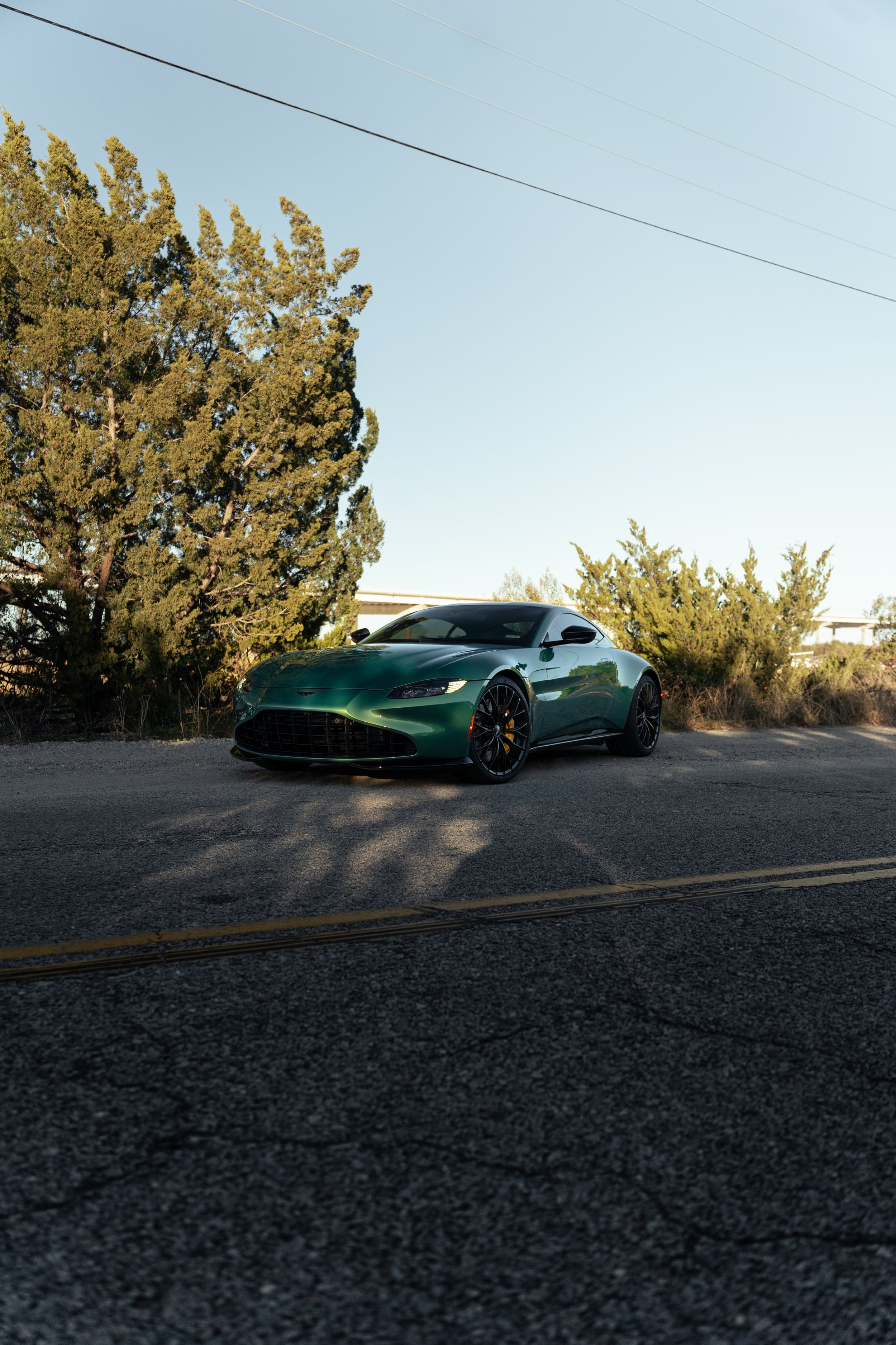 A dark green sports car parked on the side of a road with trees and a light blue sky in the background.