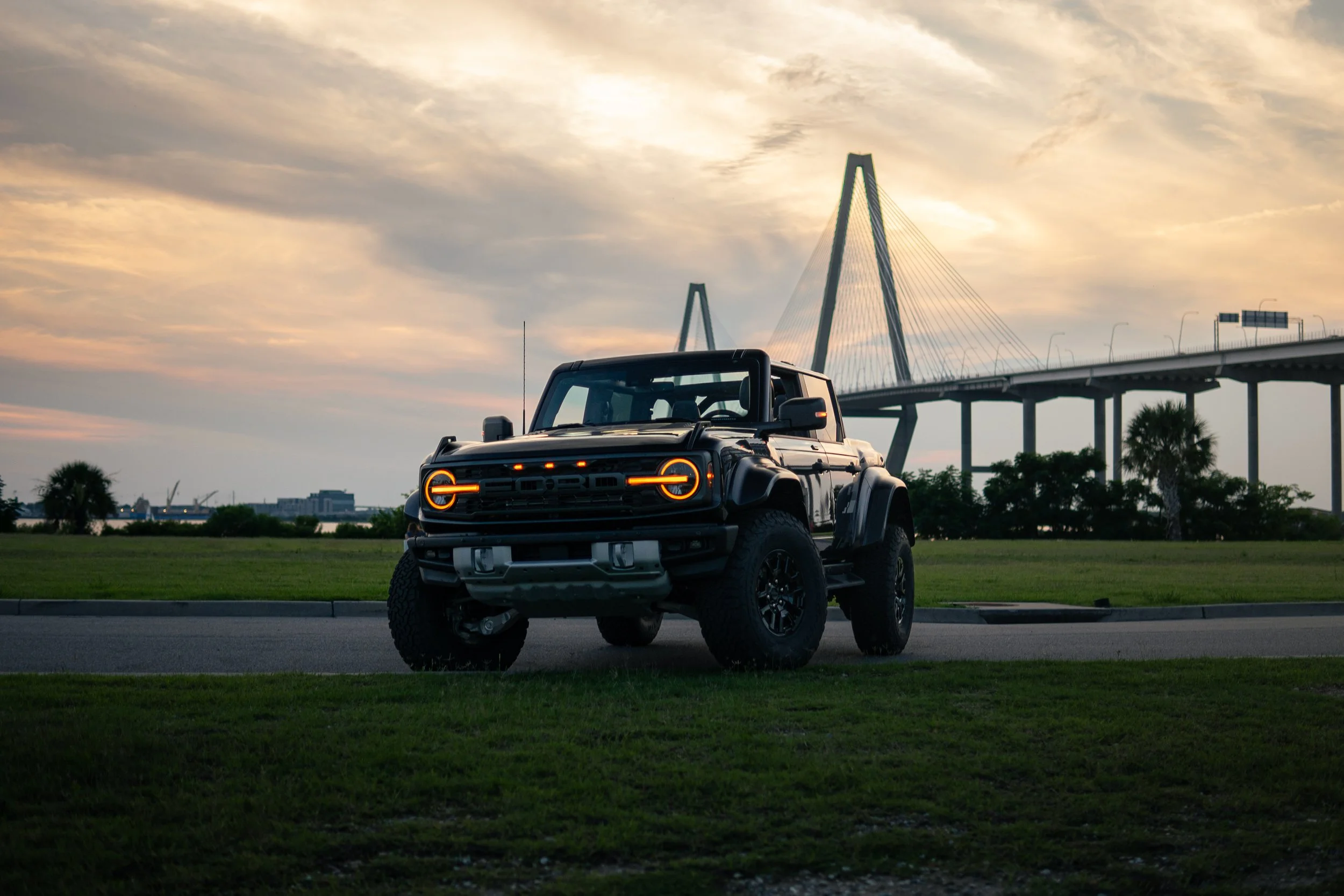 A black off-road vehicle with illuminated orange headlights parked on grass near a road, with a bridge and palm trees in the background during sunset.