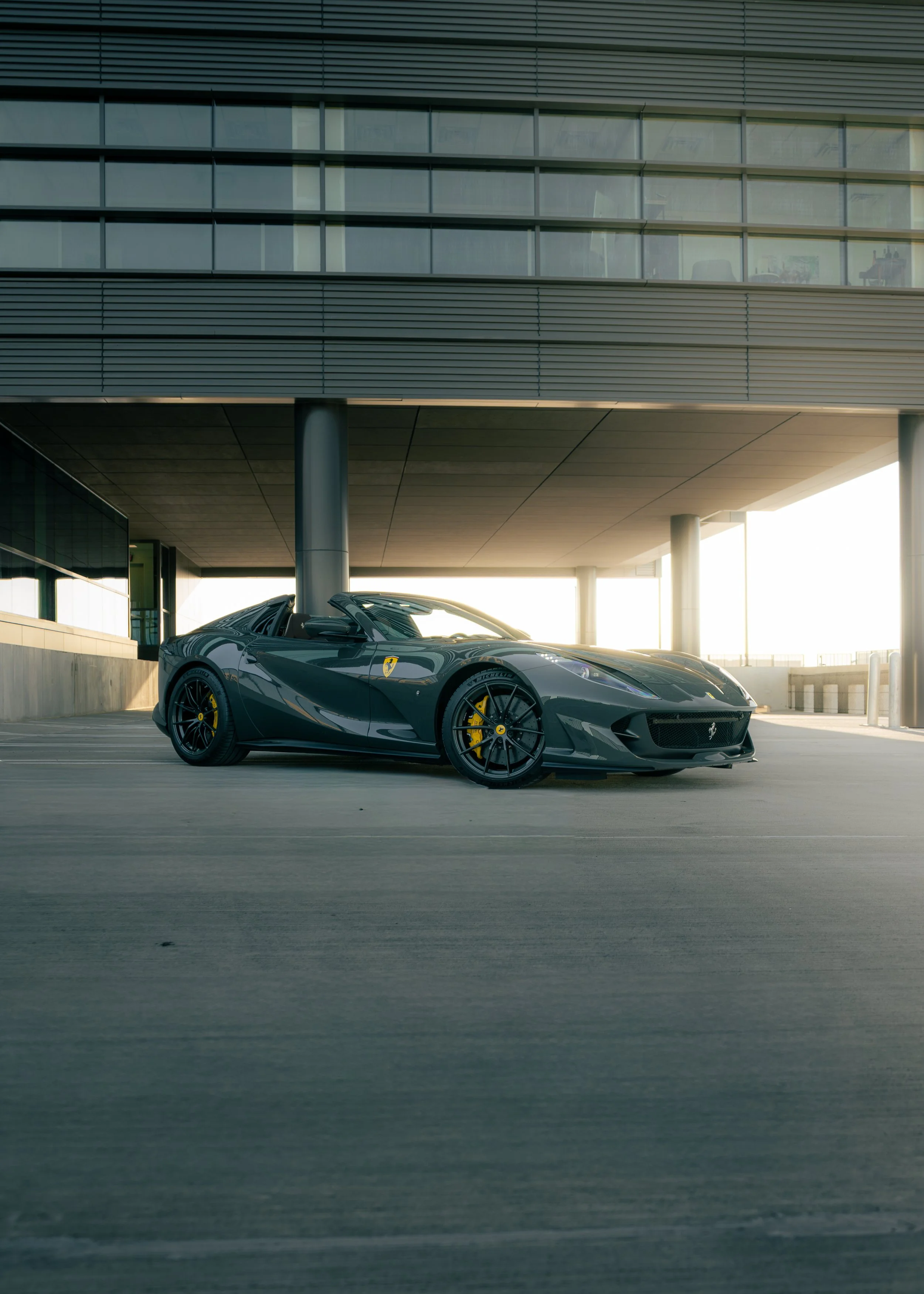 A black Ferrari sports car parked under a modern building. The car features yellow brake calipers and a sleek design, with sunlight illuminating the background.