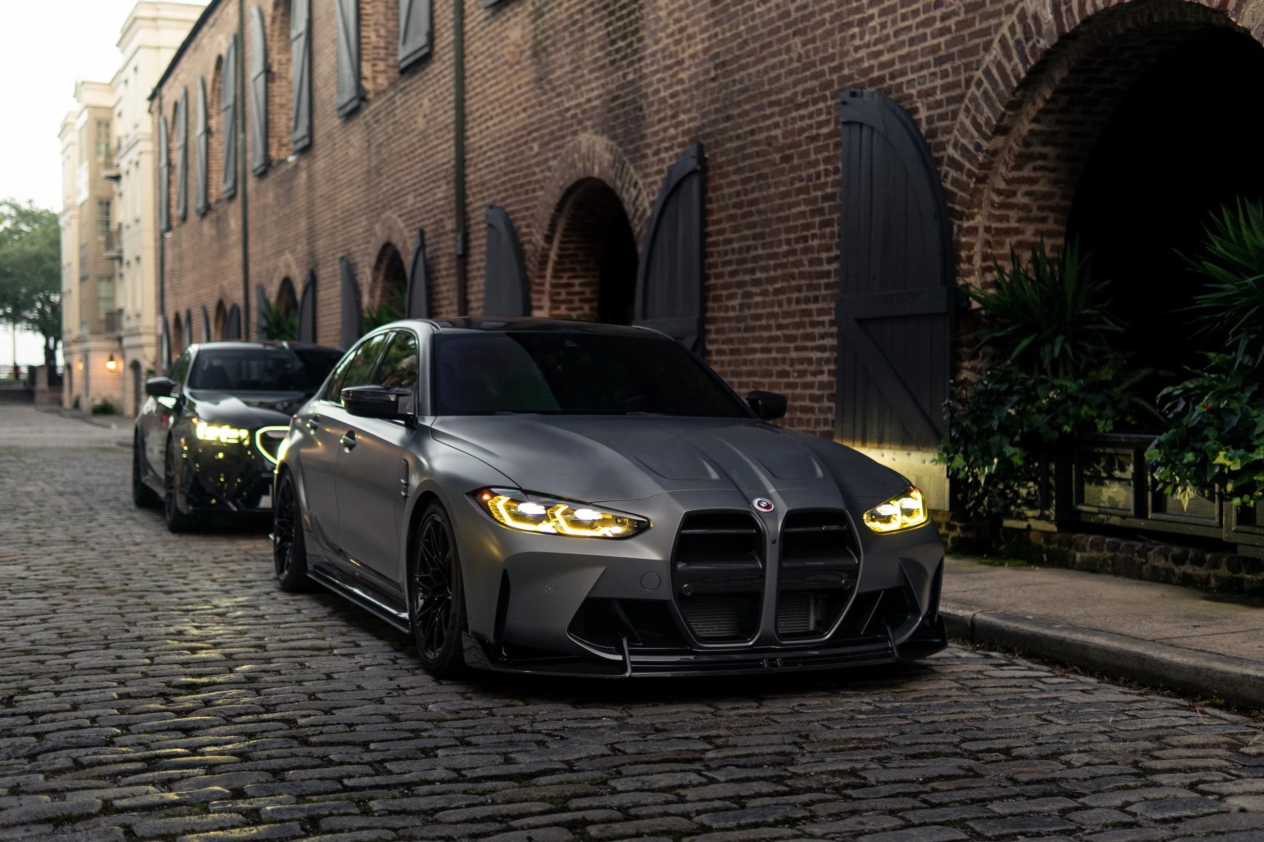 A lineup of three cars parked on a cobblestone street in front of brick buildings with arched windows during dusk. The front car is a gray BMW with yellow headlights.