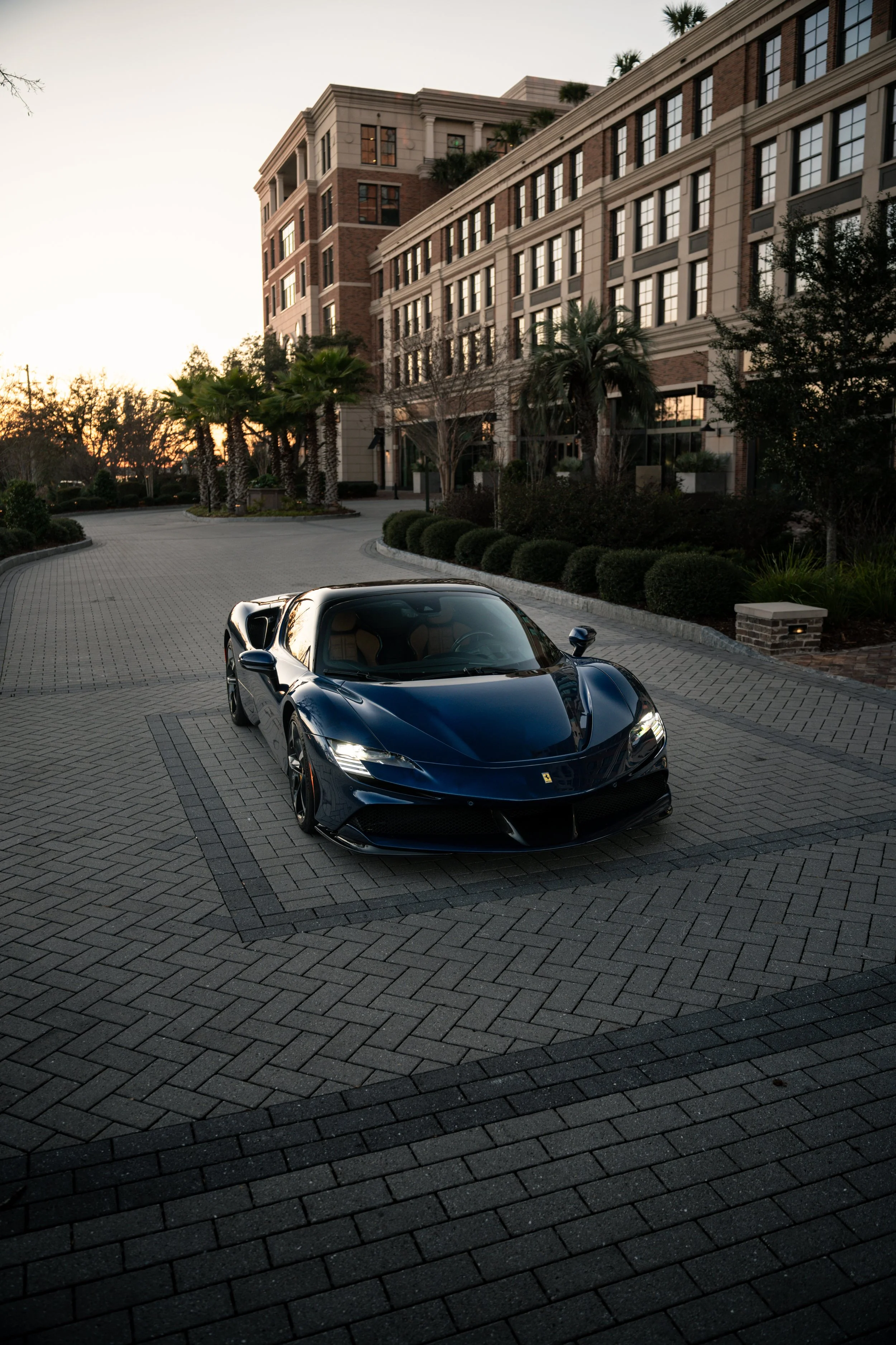 A blue sports car parked on a paved driveway in front of a large building with multiple windows and palm trees, during sunset or early evening.