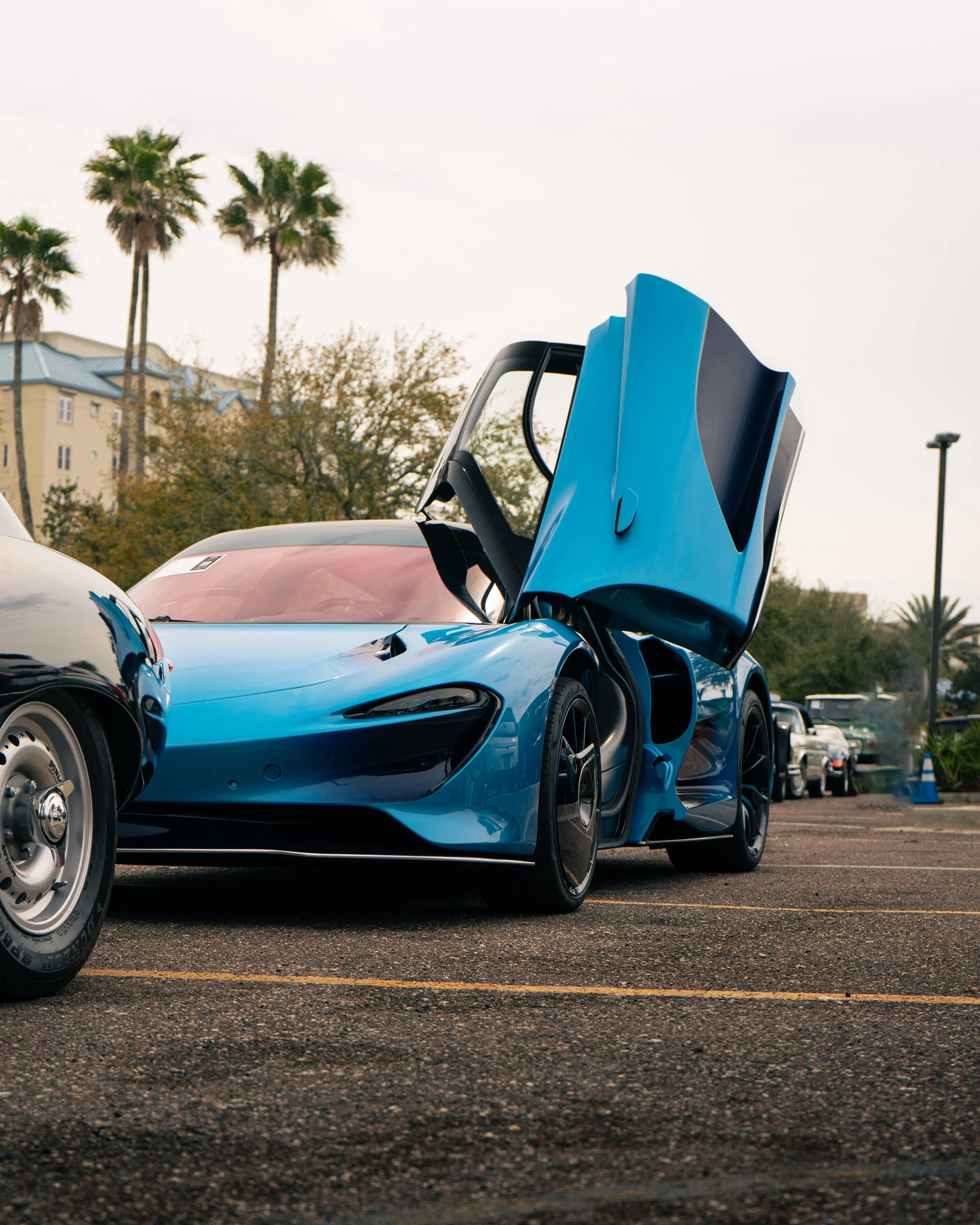A blue sports car with its gull-wing door open parked in a parking lot, with palm trees and buildings in the background.