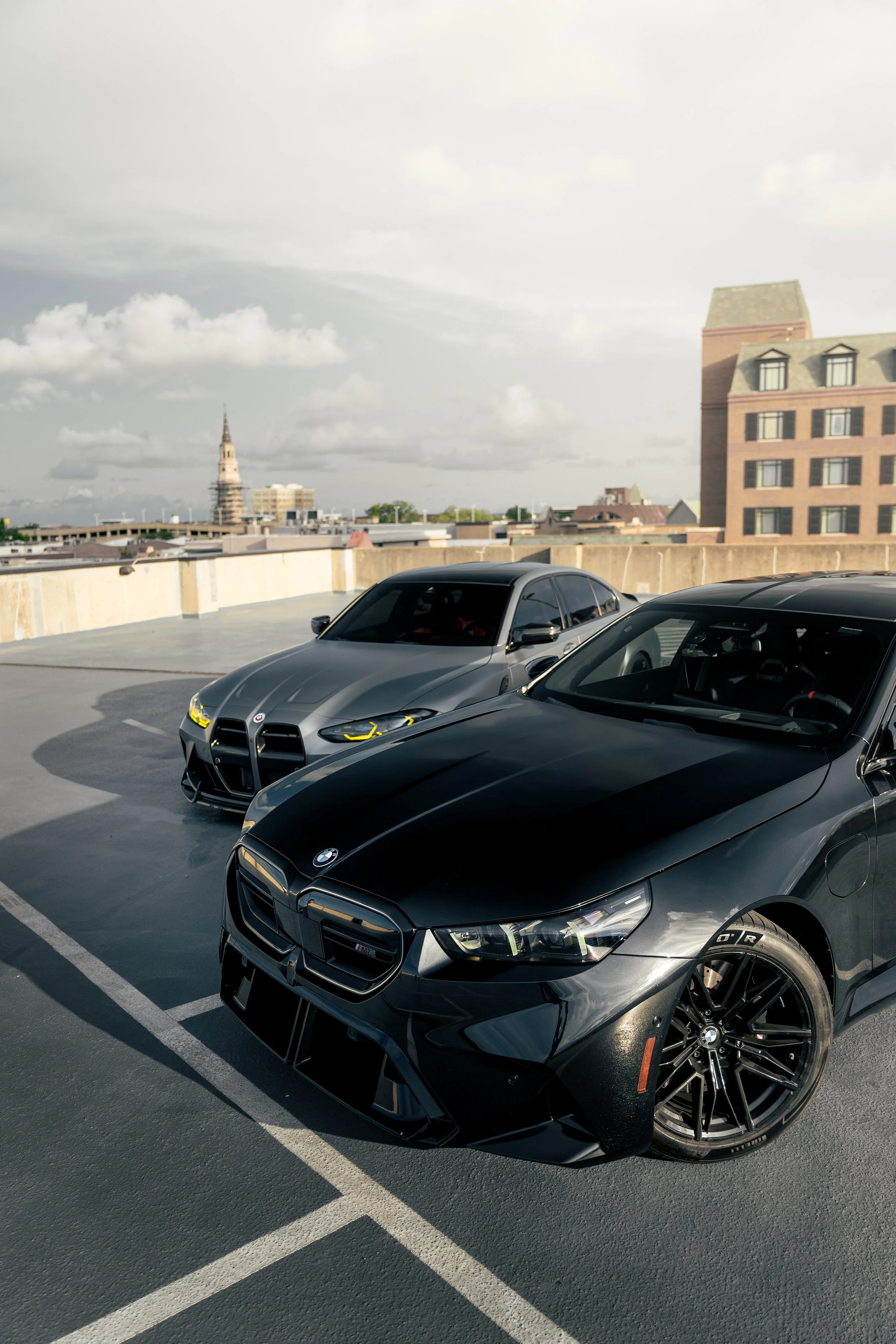 Two parked luxury cars, a black BMW and a gray Alfa Romeo, on a rooftop parking lot with city buildings and a cloudy sky in the background.
