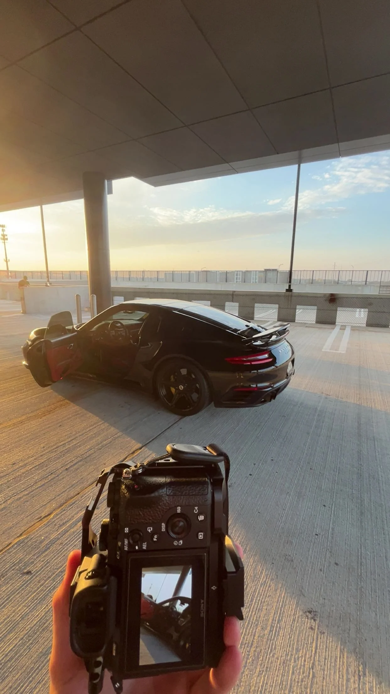 A person holding a camera taking a photo of a black sports car parked under a bridge or overpass, with a sunset sky in the background.