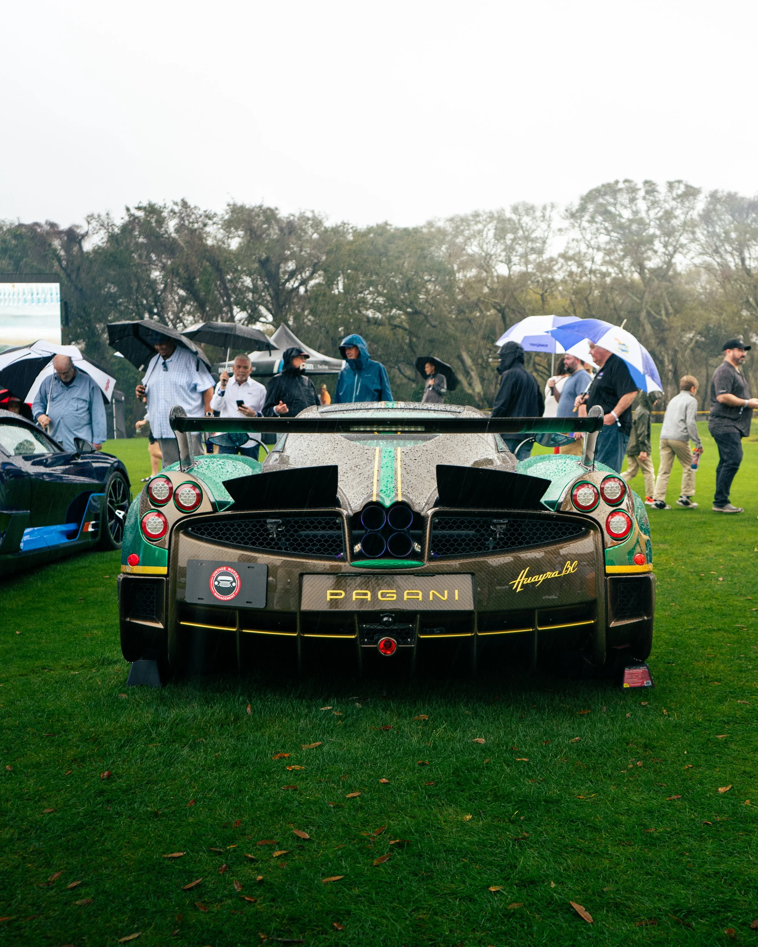 Rear view of a Pagani Huayra BC supercar on display at an outdoor event, with people holding umbrellas in the background.