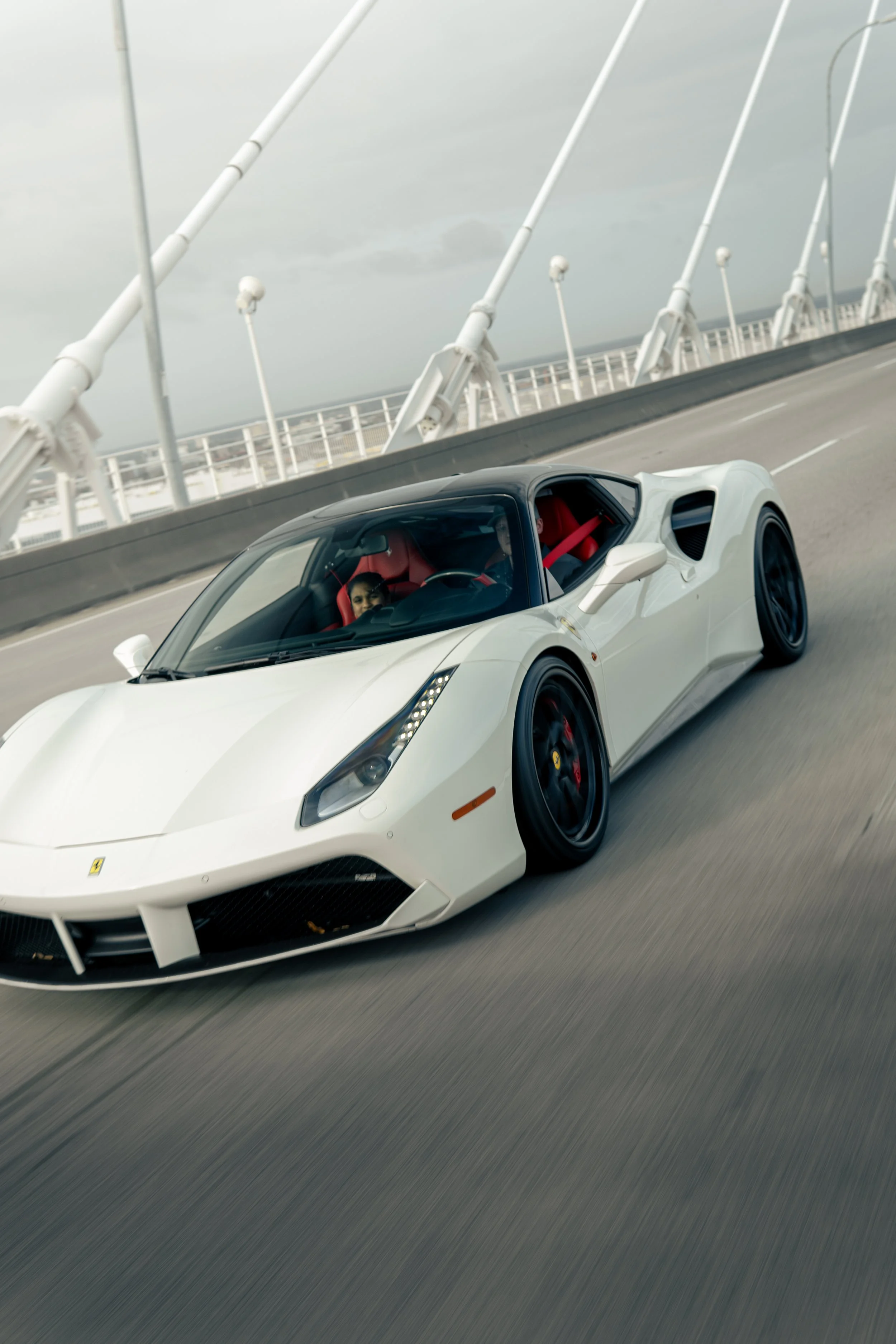 A white convertible sports car, a Ferrari, driving on a bridge with a cloudy sky above.
