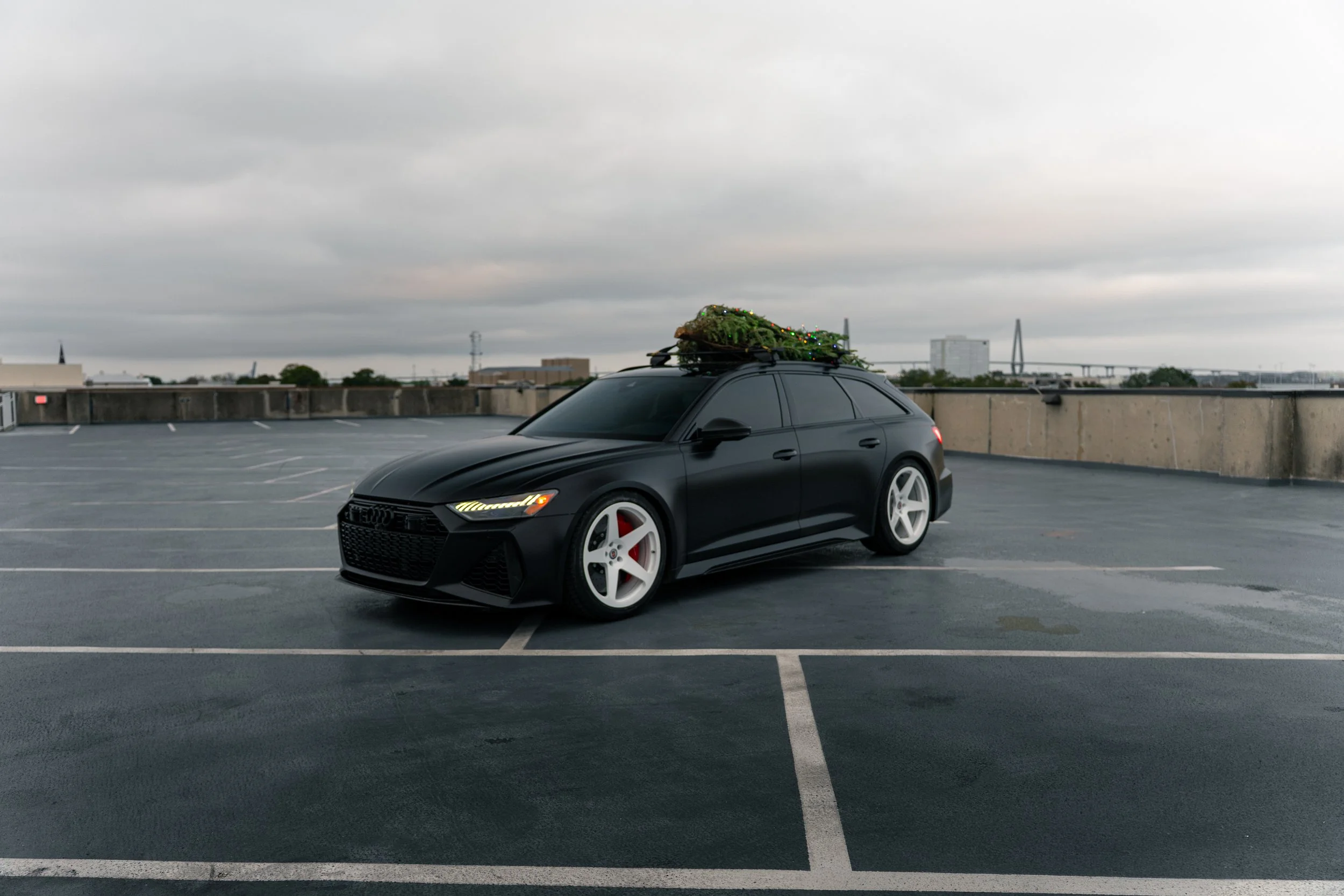 Black Audi station wagon parked in an empty rooftop parking lot with a Christmas tree on its roof rack.