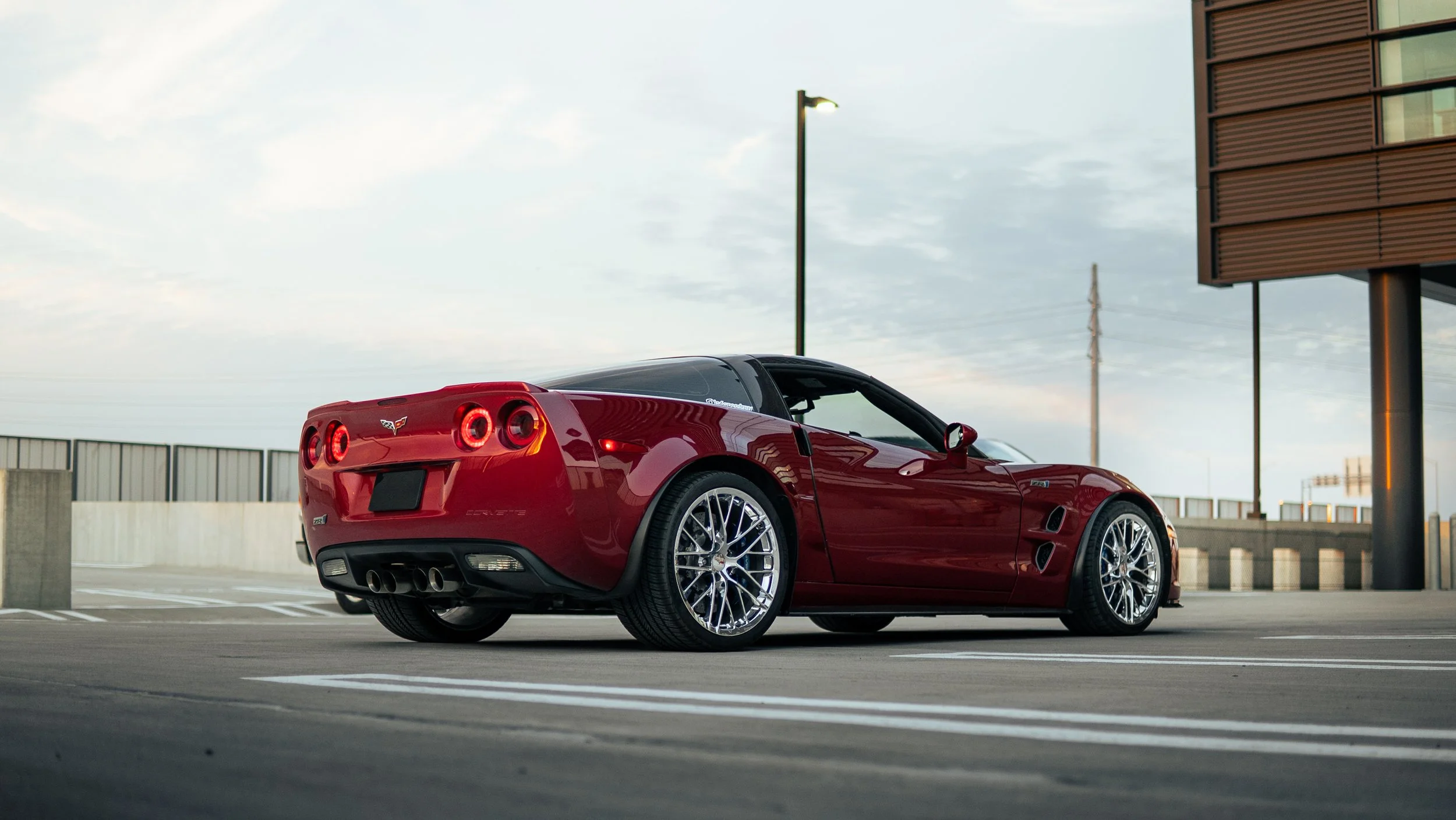 Red Chevrolet Corvette sports car parked in an empty rooftop parking lot with a cityscape background and cloudy sky.
