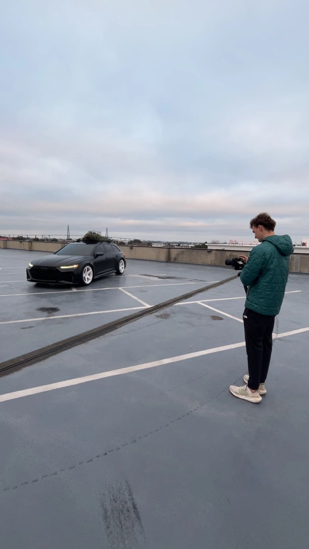 A person filming a black car with a camera on a rooftop parking lot during overcast weather.