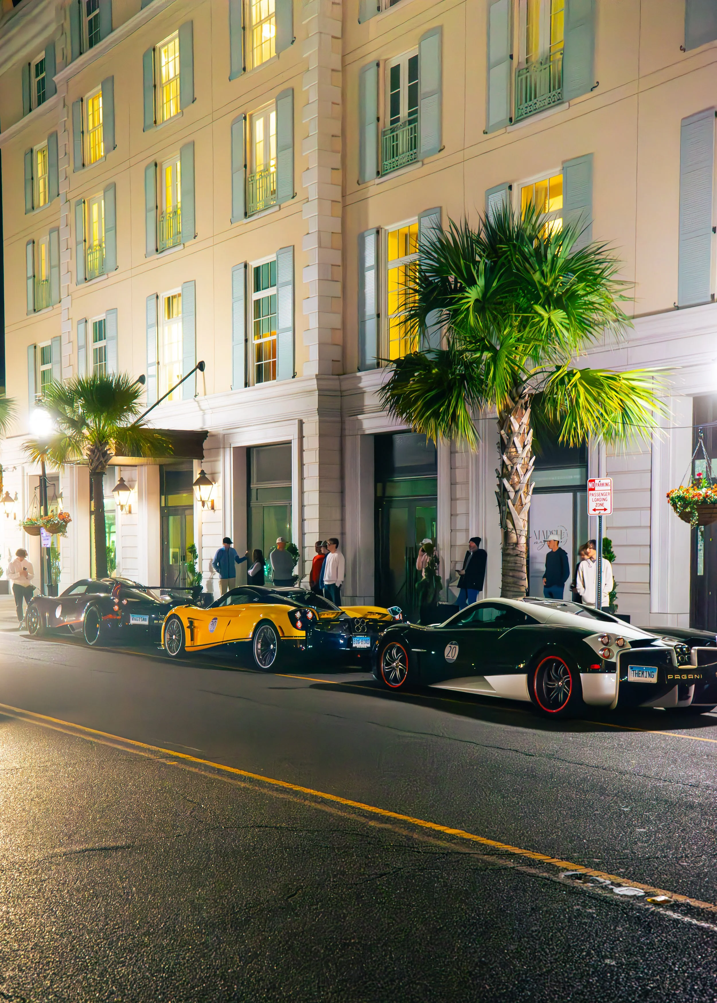 Night scene on a city street with three luxury sports cars parked along the curb. The building behind features tall windows with green shutters and palm trees are visible on sidewalk. Pedestrians are seen walking and standing near the entrance.