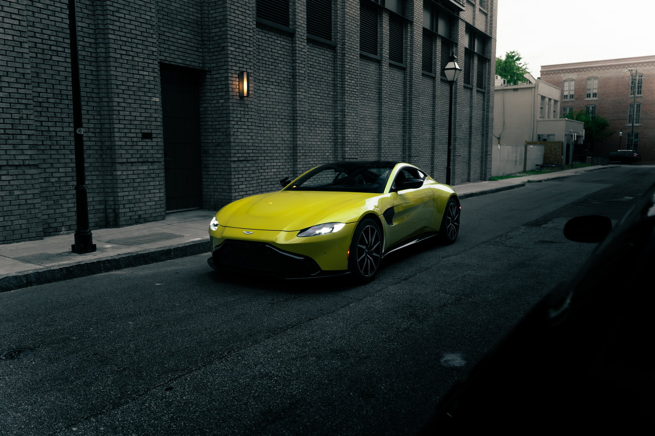 A bright yellow sports car parked on a city street next to a gray brick building with a black door and street lamps.