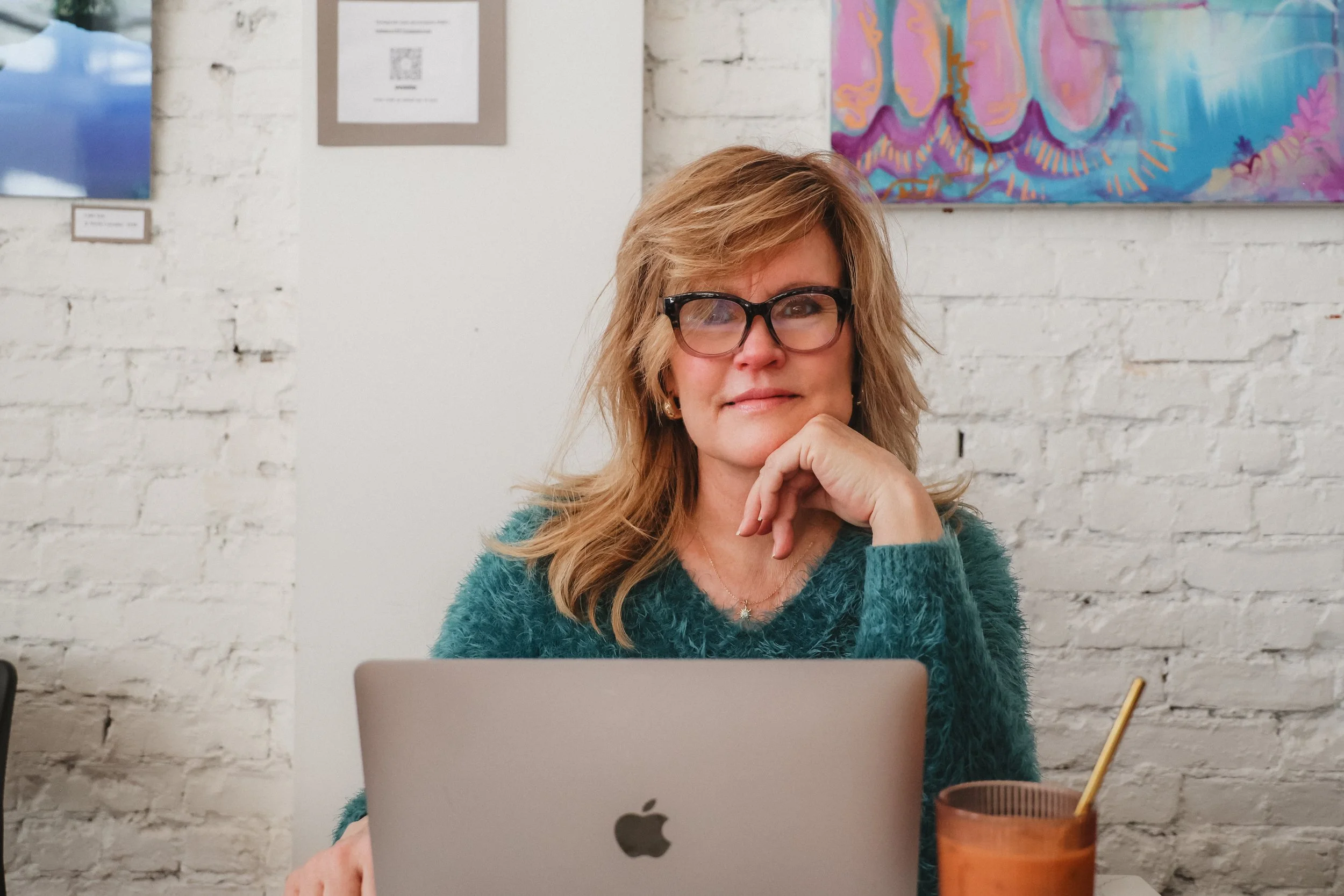 A woman with wavy blonde hair and glasses sitting at a table in front of a silver MacBook, resting her chin on her hand, in a room with white brick walls and colorful artwork.