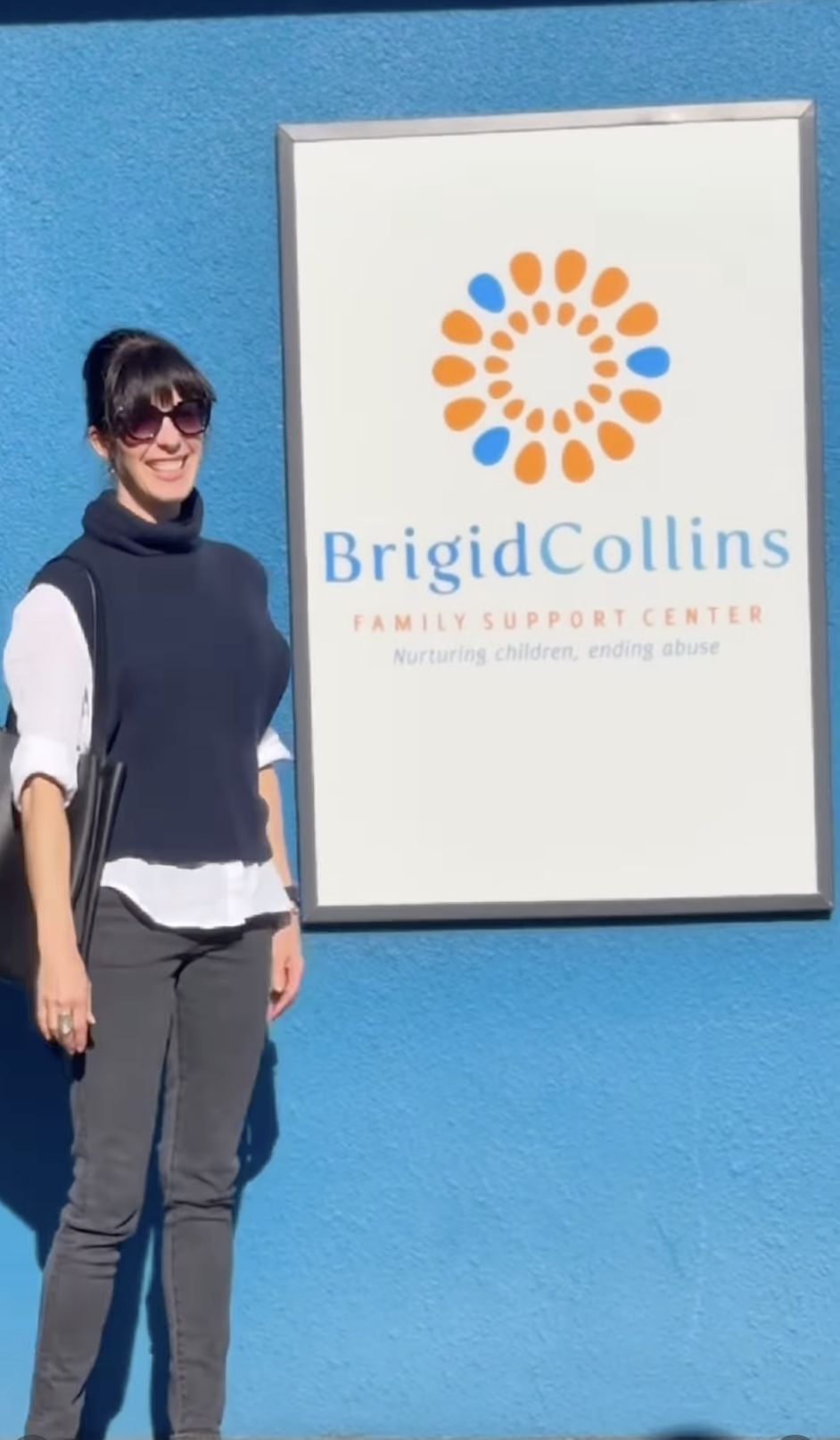 A woman standing outside next to a sign for Brigid Collins Family Support Center, with a blue wall background. The sign has a circular logo in orange and blue, and the text 'Brigid Collins Family Support Center' with a tagline 'Nurturing children, ending abuse'.