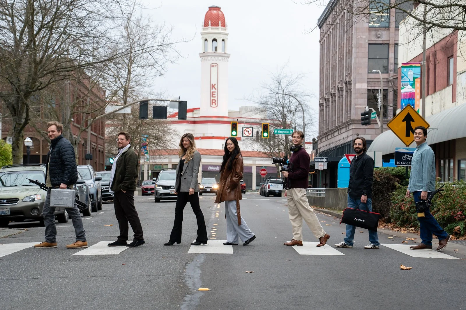 A group of seven people crossing a city street at a crosswalk, with a vintage theater building in the background and cars parked along the sides of the street.