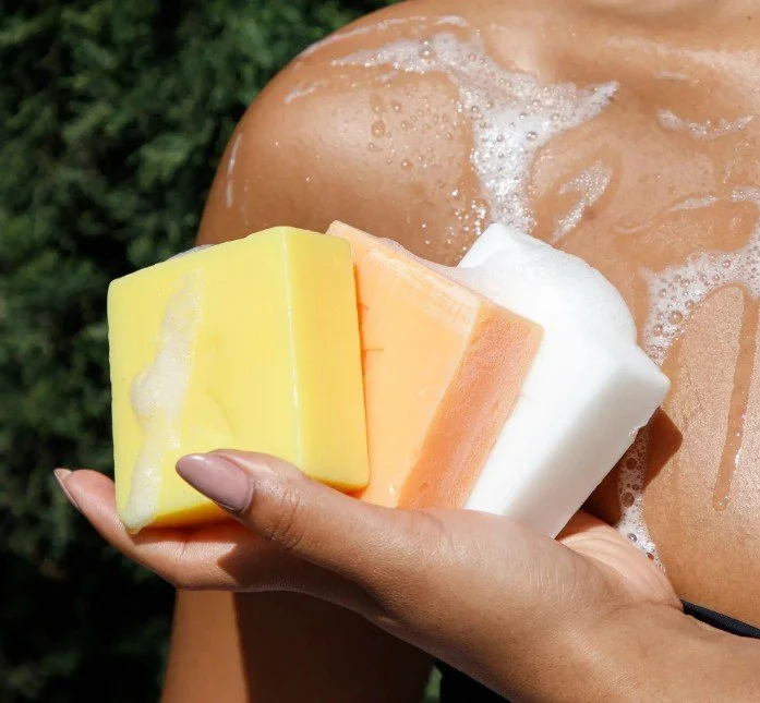A person holding three bars of soap in front of their shoulder, with a background of green foliage and soap bubbles on their skin.