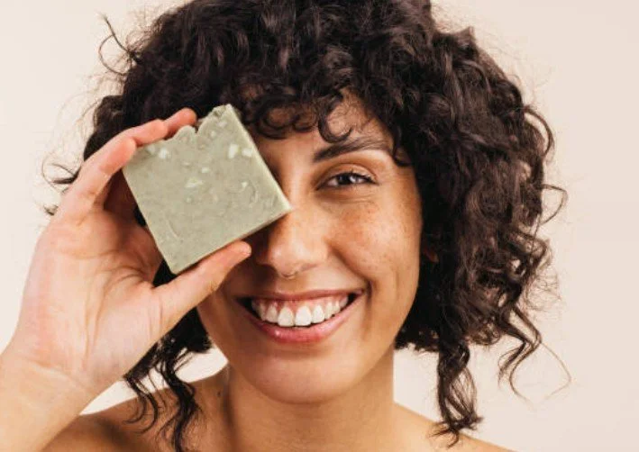 A woman with curly hair smiling and holding a bar of soap over one eye.