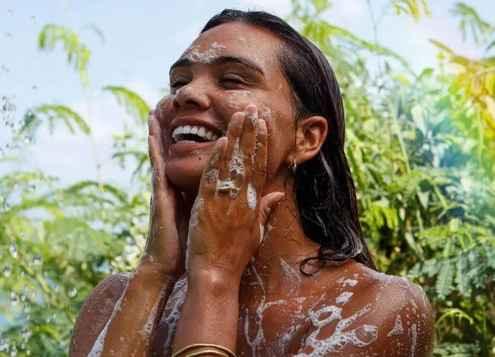 A woman with wet hair and a joyful expression washing her face outdoors with soap and water, surrounded by greenery.
