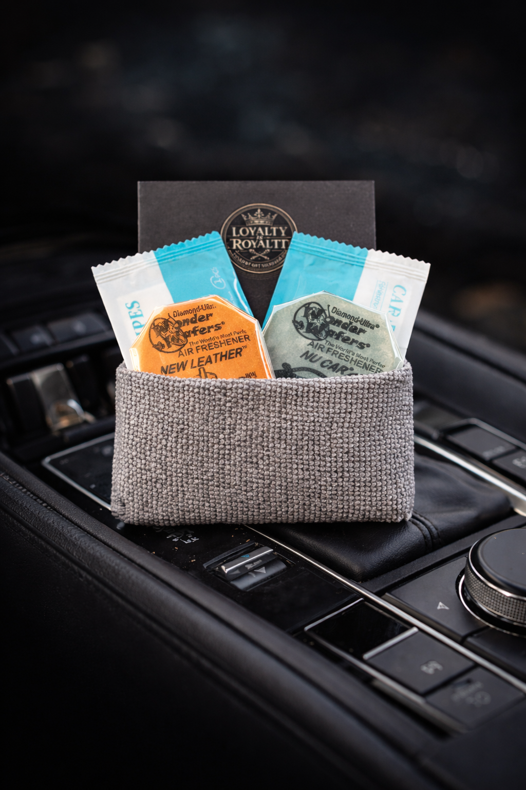 Car interior with a small grey fabric pouch containing two air freshener packets, one orange labeled 'New Leather' and one green labeled 'Nu Car', placed on the center console.
