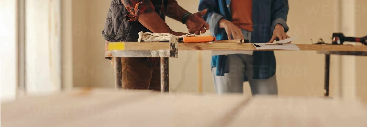 Two people are working on a construction project, standing at a table with tools and papers.