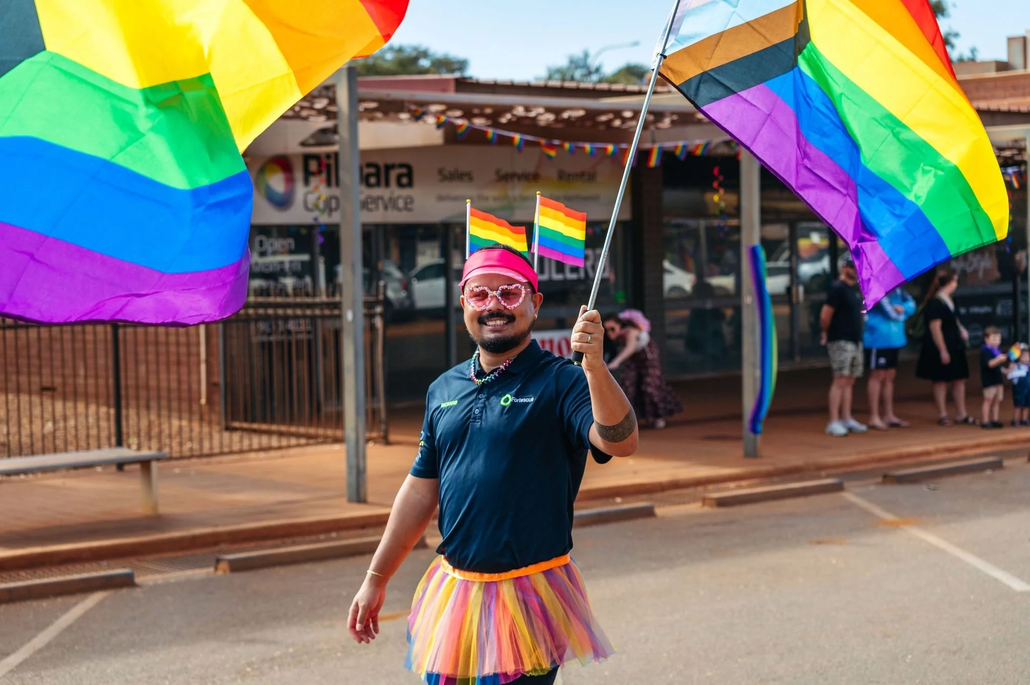 A person smiling at a pride parade, holding rainbow flags, dressed in a rainbow skirt, pink sunglasses, and a pink headband, surrounded by others with pride decorations.