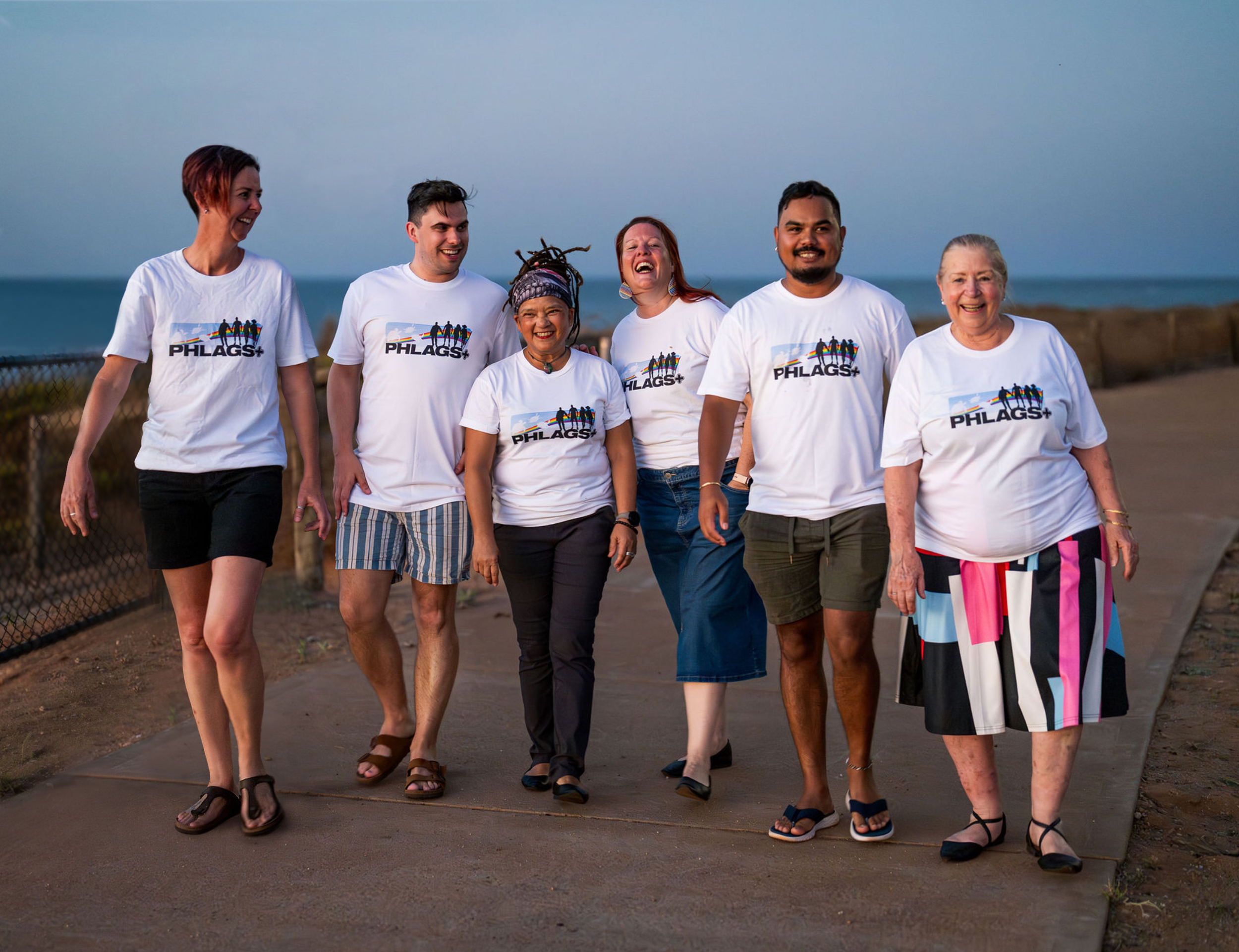 Group of diverse people walking together outdoors near the beach, all wearing white t-shirts with the 'PHLAGS+' logo, smiling and enjoying each other's company.