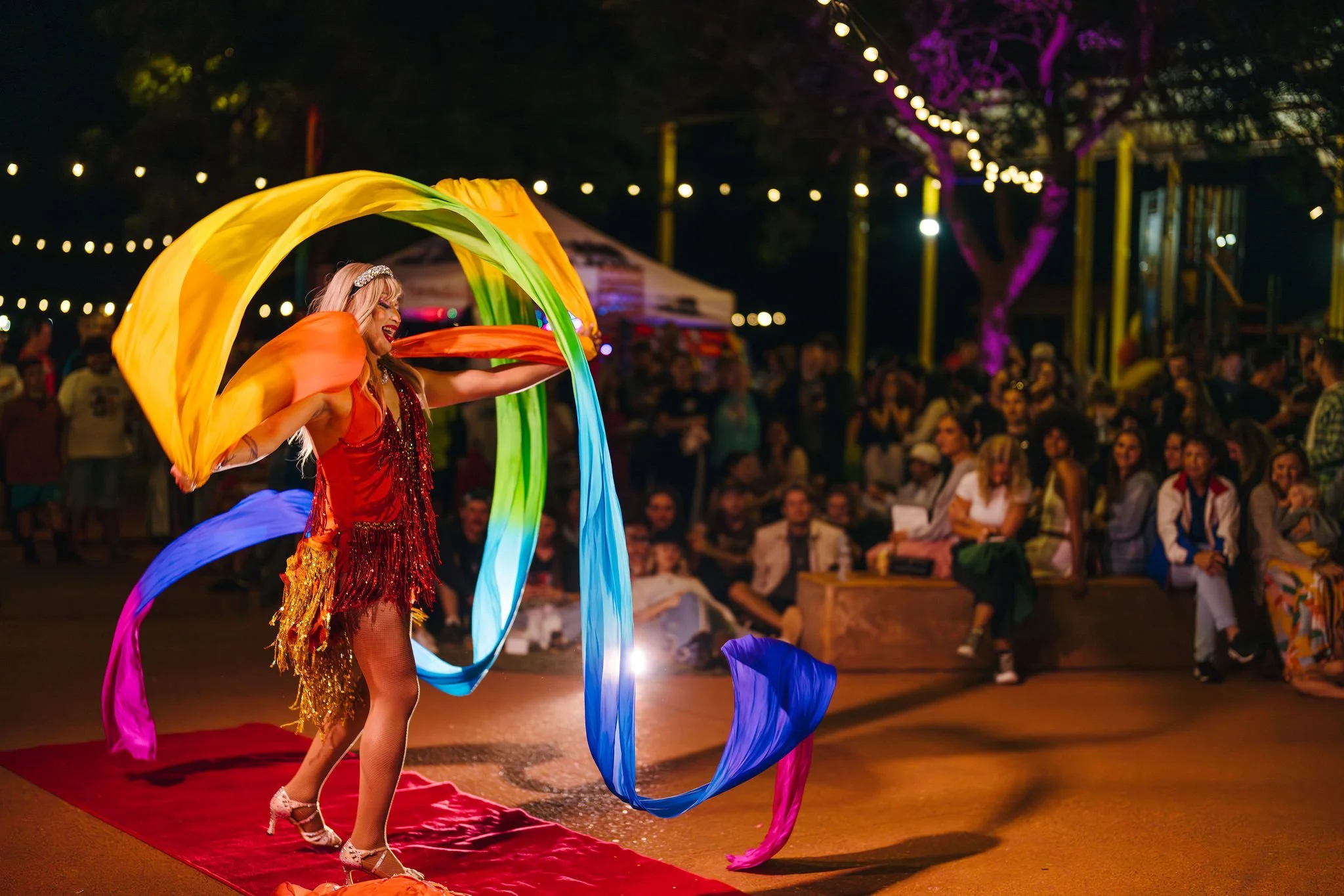 A woman performing a ribbon dance on a red carpet at night, with a crowd of spectators watching. She is dressed in a red, fringed costume and is holding colorful ribbons, with string lights and purple-lit trees in the background.