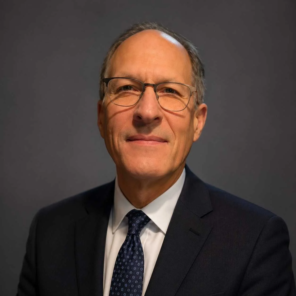 A middle-aged man wearing glasses, a dark suit, white shirt, and patterned tie, posing against a plain dark background.