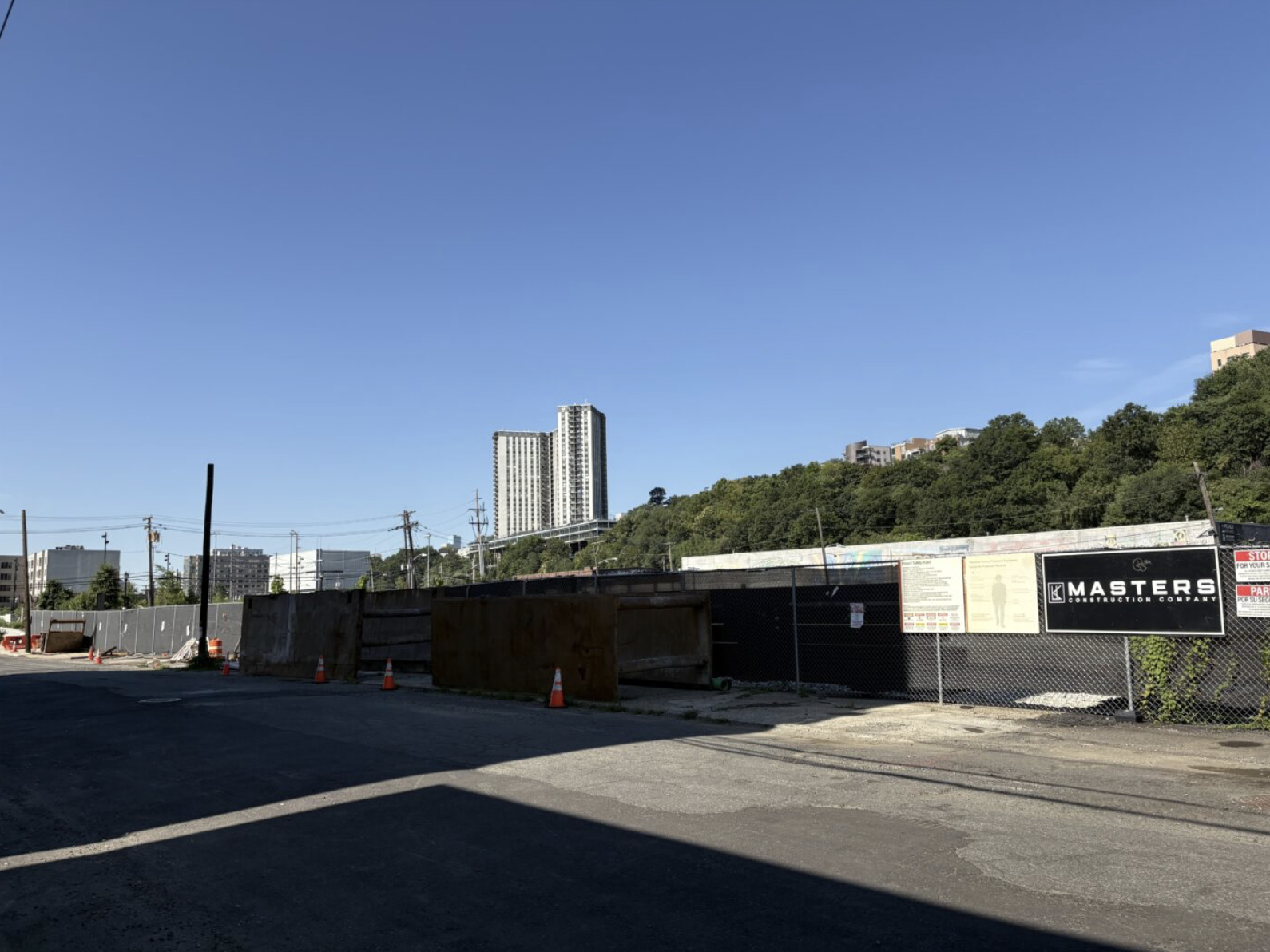 Construction site with a fence, orange cones, and signs, with a cityscape and green hillside in the background under a clear blue sky.