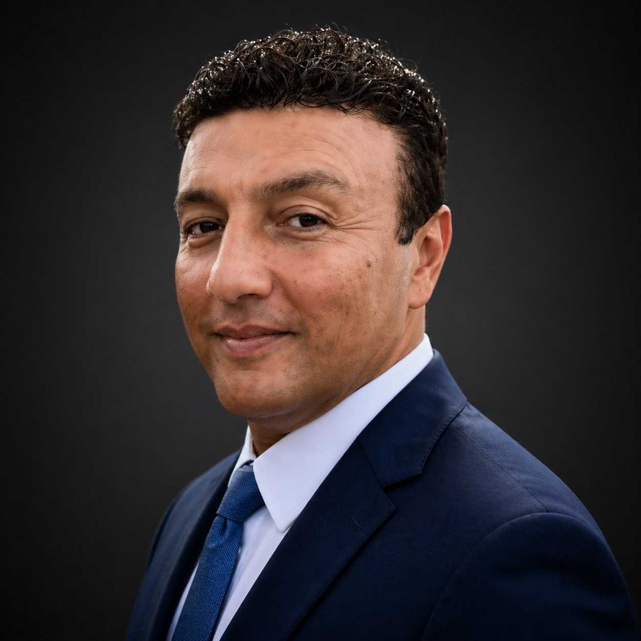 Headshot of a man wearing a navy suit, white shirt, and blue tie, with a confident expression, short dark curly hair, and slight smile, set against a dark background.