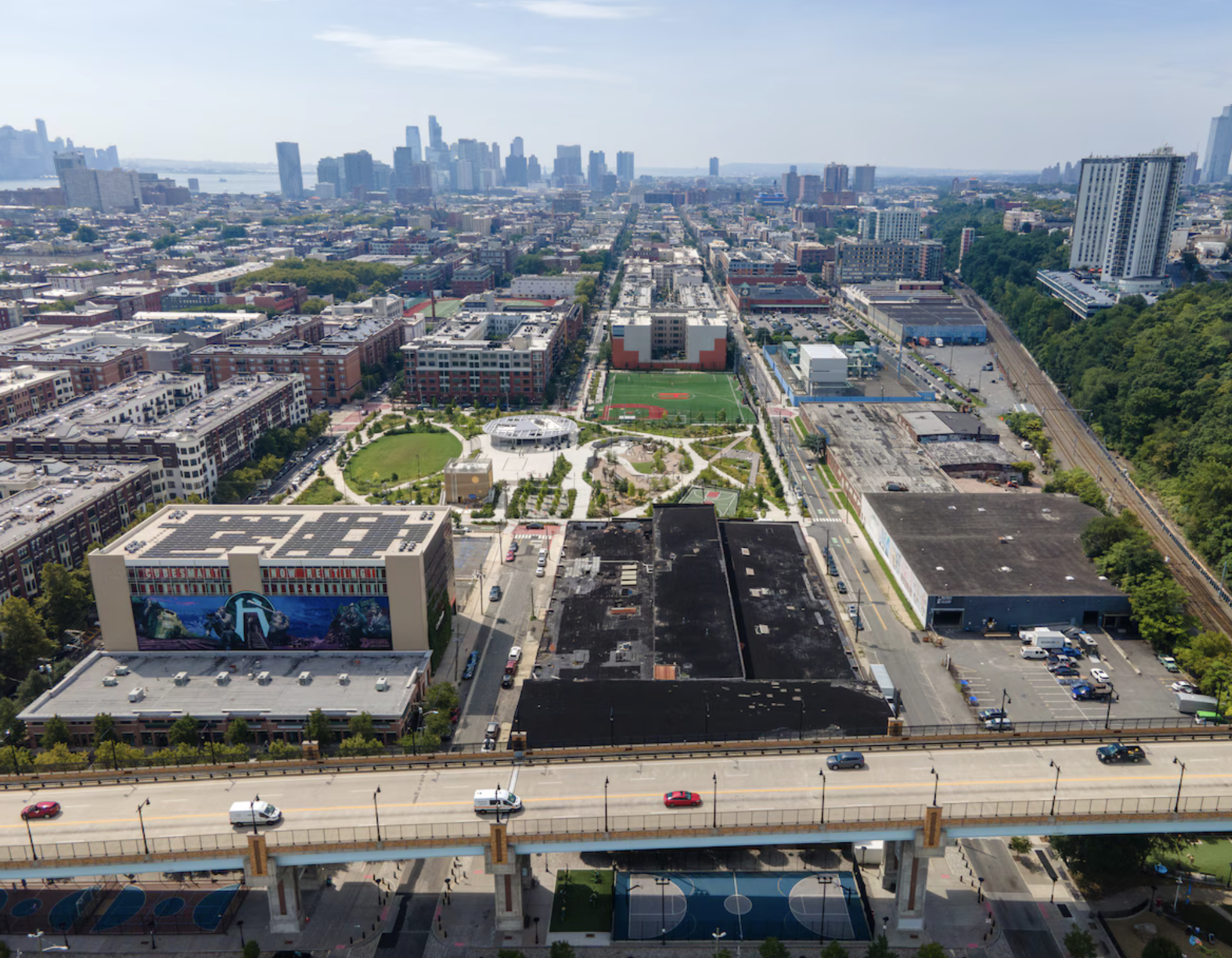 Aerial view of a city skyline with tall buildings, a river, and a mix of residential and commercial areas. There are sports fields, parking lots, and train tracks visible in the foreground. A highway runs across the lower part of the image.