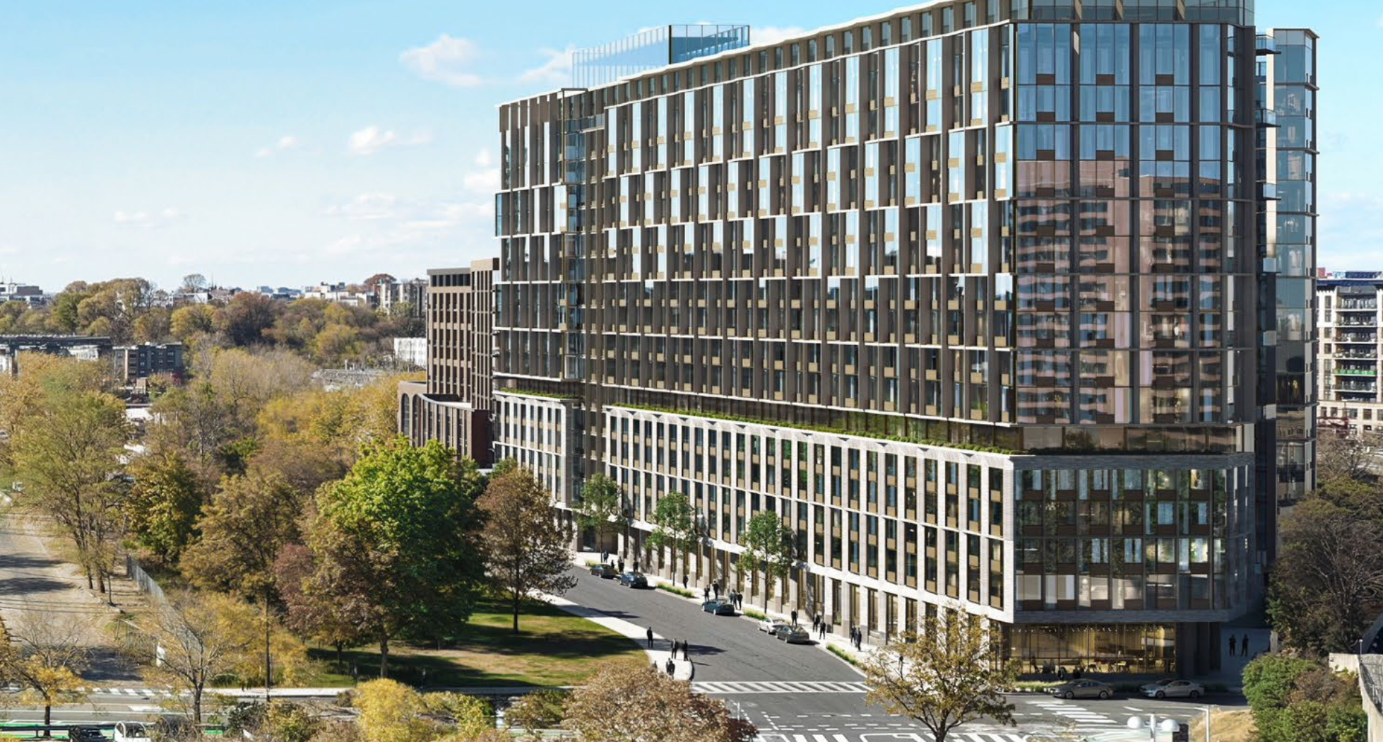 Modern multi-story glass building with trees and a cityscape in the background on a clear day.