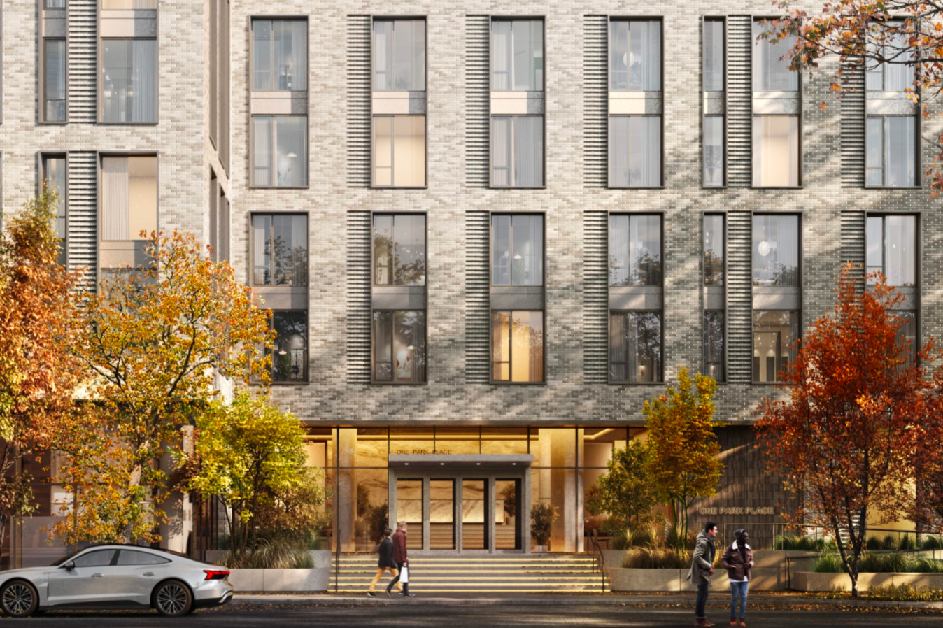 Modern brick apartment building with large windows and fall foliage in front, with two pedestrians walking and a parked car on the street.