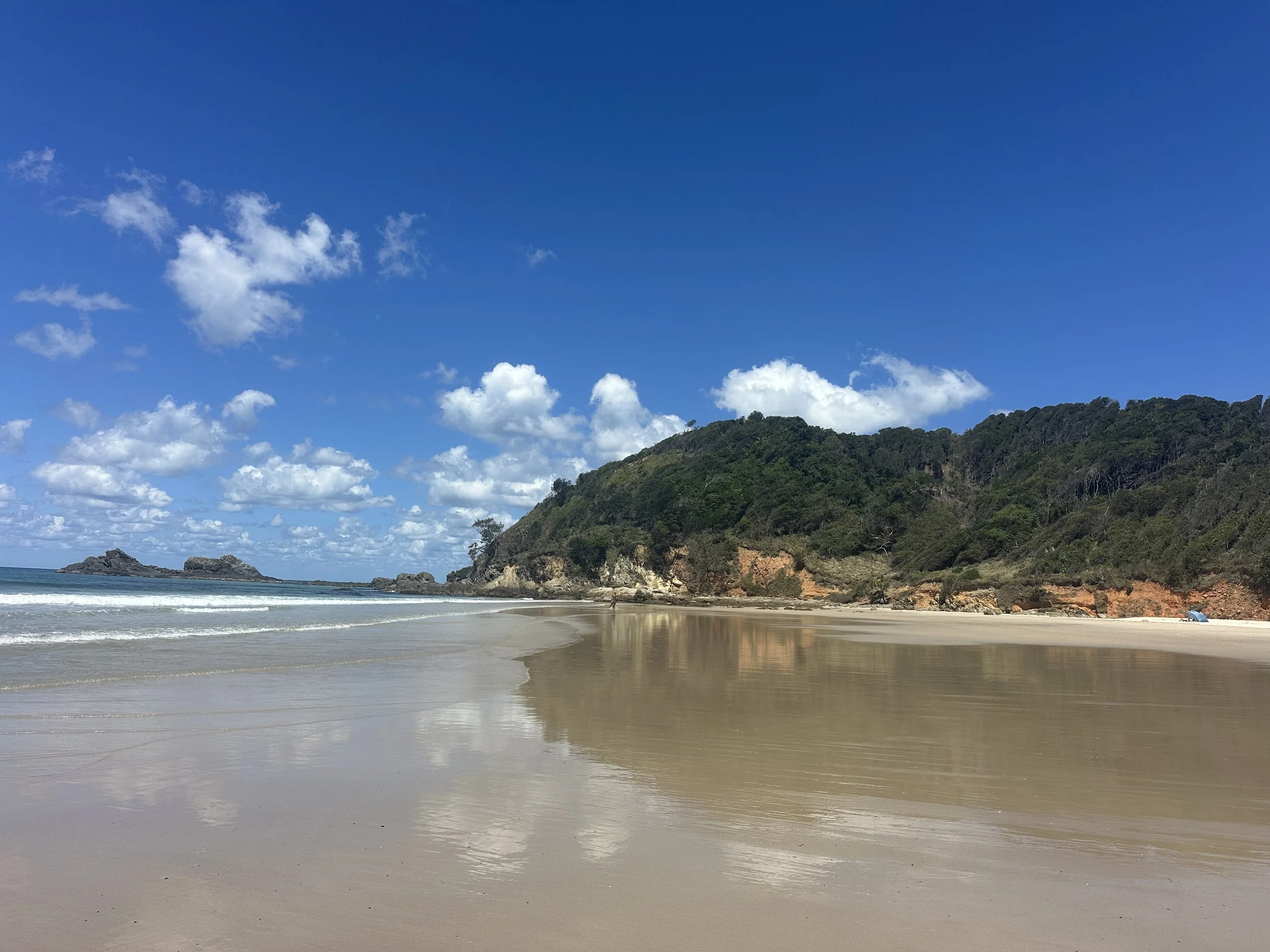 A scenic beach with a sandy shore, blue sky with white clouds, green hillside, and calm ocean waves.