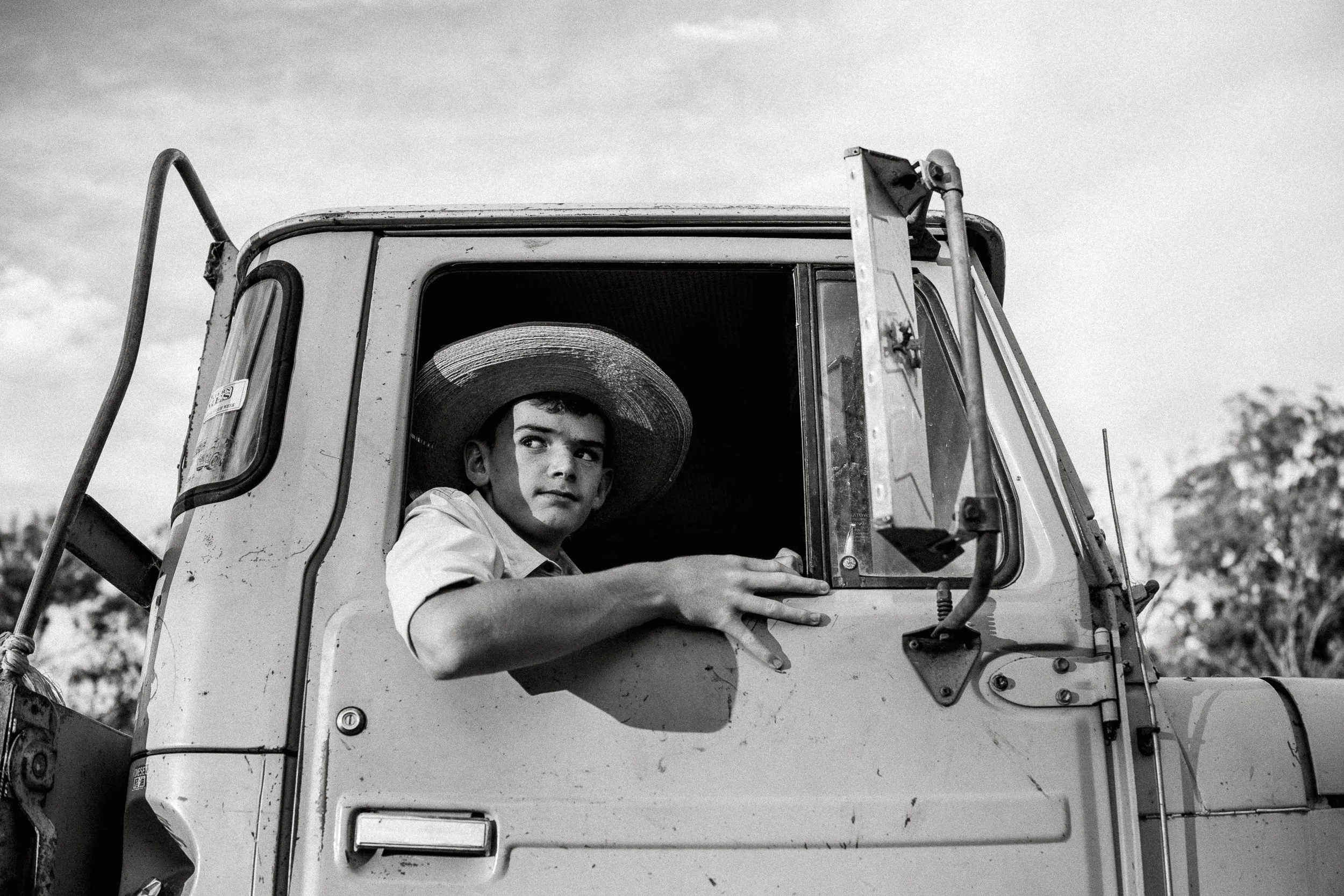 A boy with a straw hat sitting in the driver's seat of an old tractor, looking out the window.