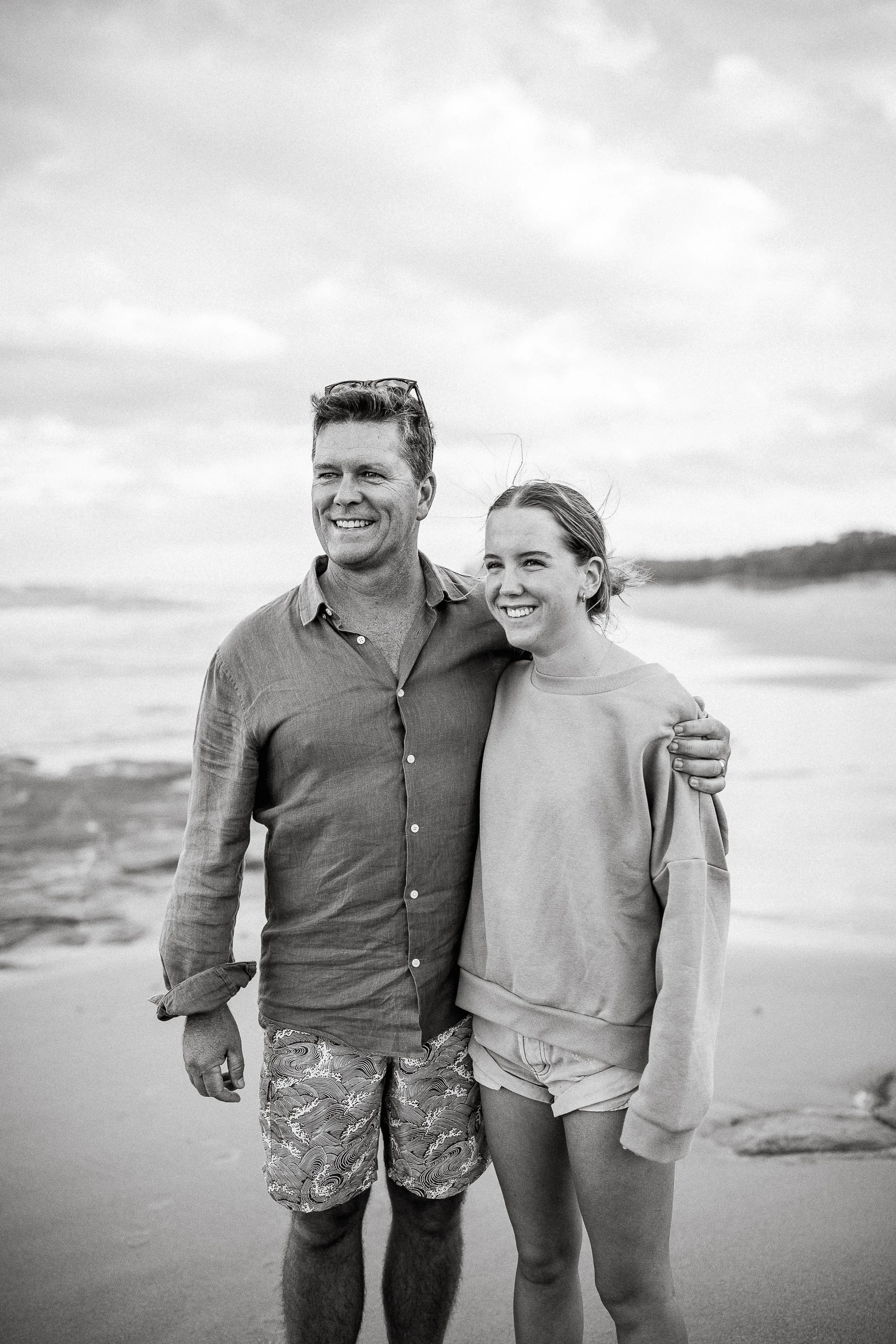 A father and a daughter standing arm in arm on the beach, smiling, with the ocean and sky in the background, in black and white.