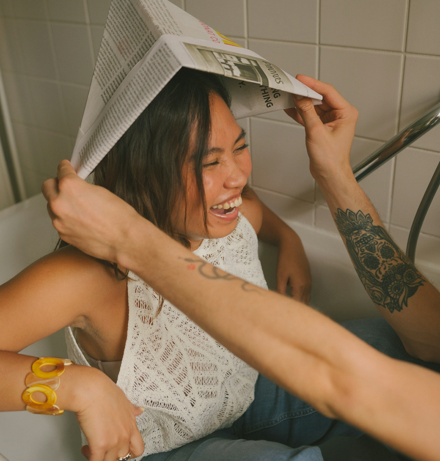 A woman in a white sleeveless top laughing as a person with tattooed arms puts a folded newspaper on her head in a bathroom with tiled wall.