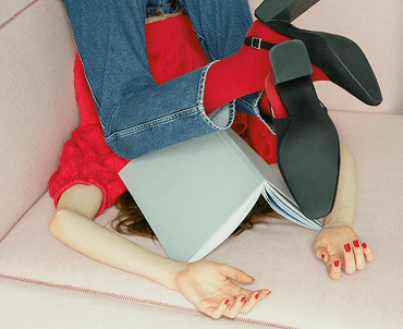 A person lying upside down on a pink sofa, wearing red boots, blue jeans, and a red sweater, with an open book resting on their stomach.