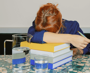 A person with red hair resting their head on a stack of books on a table, with a pitcher and glass of water nearby.