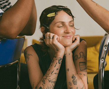A woman with short dark hair and tattoos on her arms sitting at a table, smiling with her eyes closed, resting her chin on her hands, while someone brushes her hair.