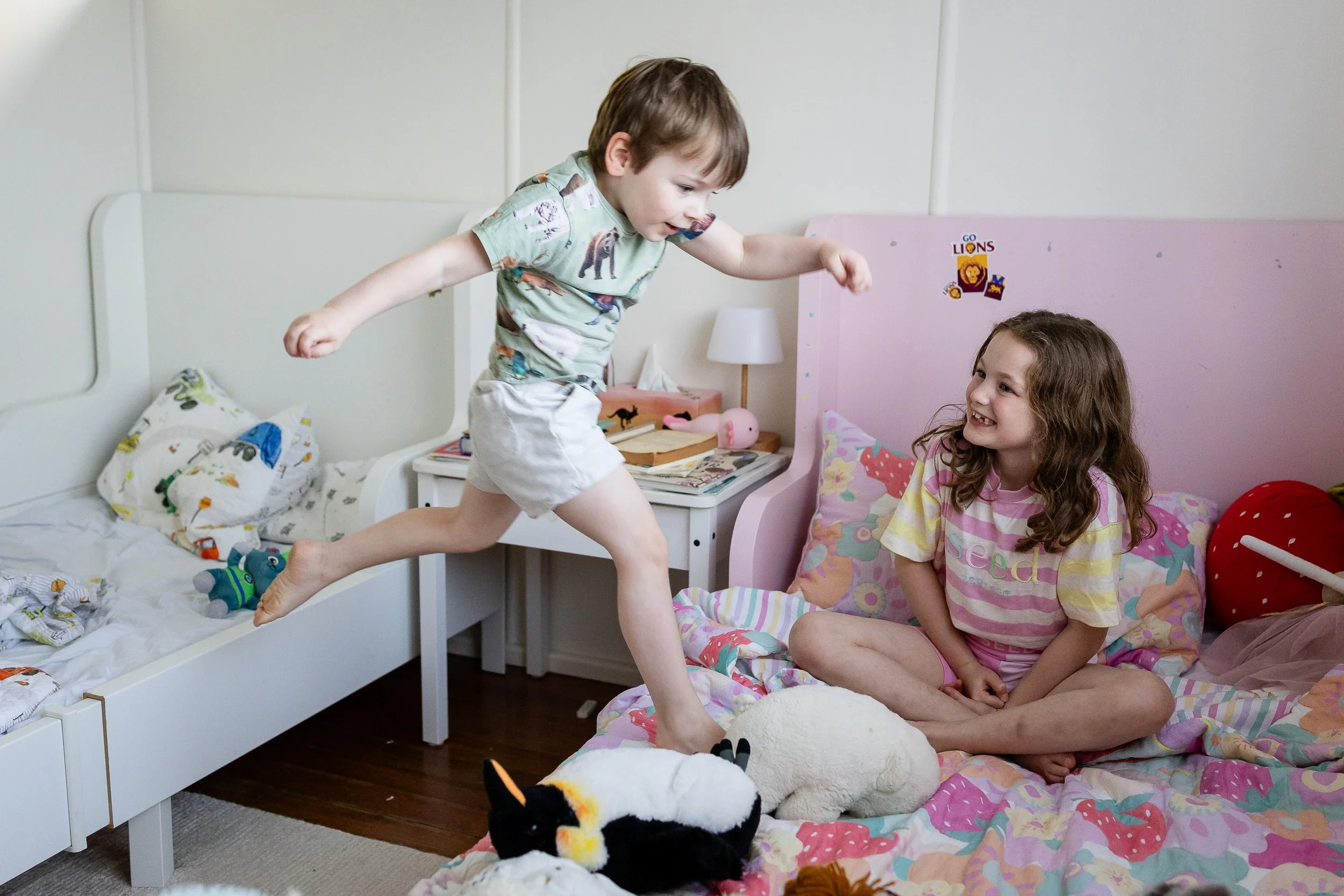 A young boy is jumping from a bed to a girl sitting cross-legged on another bed, smiling. The girl is sitting on a colorful bed with stuffed animals, including a sheep and a penguin. The room has a pink headboard, a nightstand with books and toys, and a white wall.