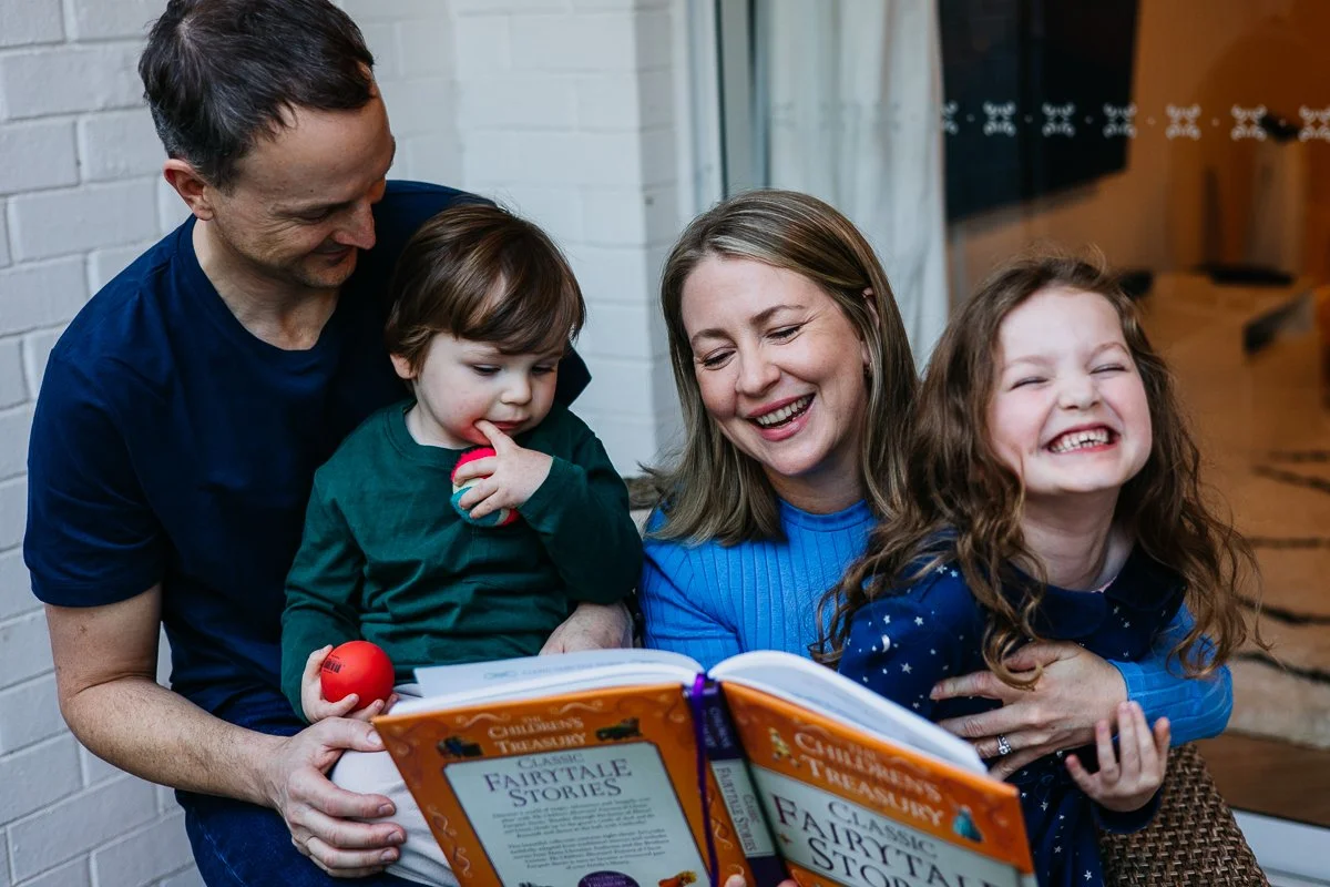 A family of four, parents with two children, sitting together reading a storybook titled 'Fairy Tale Stories', smiling and enjoying their time.