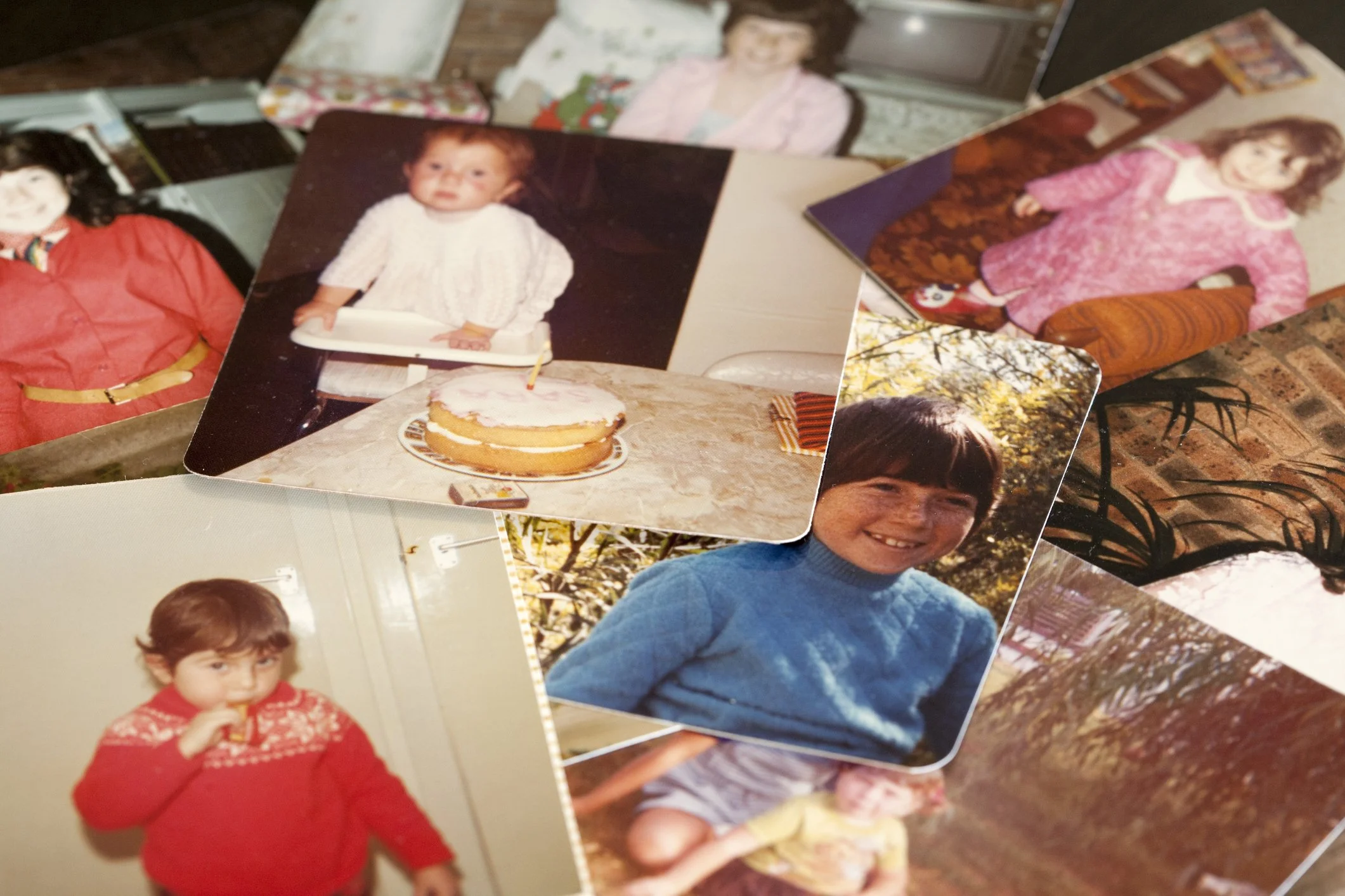 A collection of nostalgic childhood photographs scattered on a surface, including children posing indoors and outdoors, and a birthday cake with a child standing behind it.