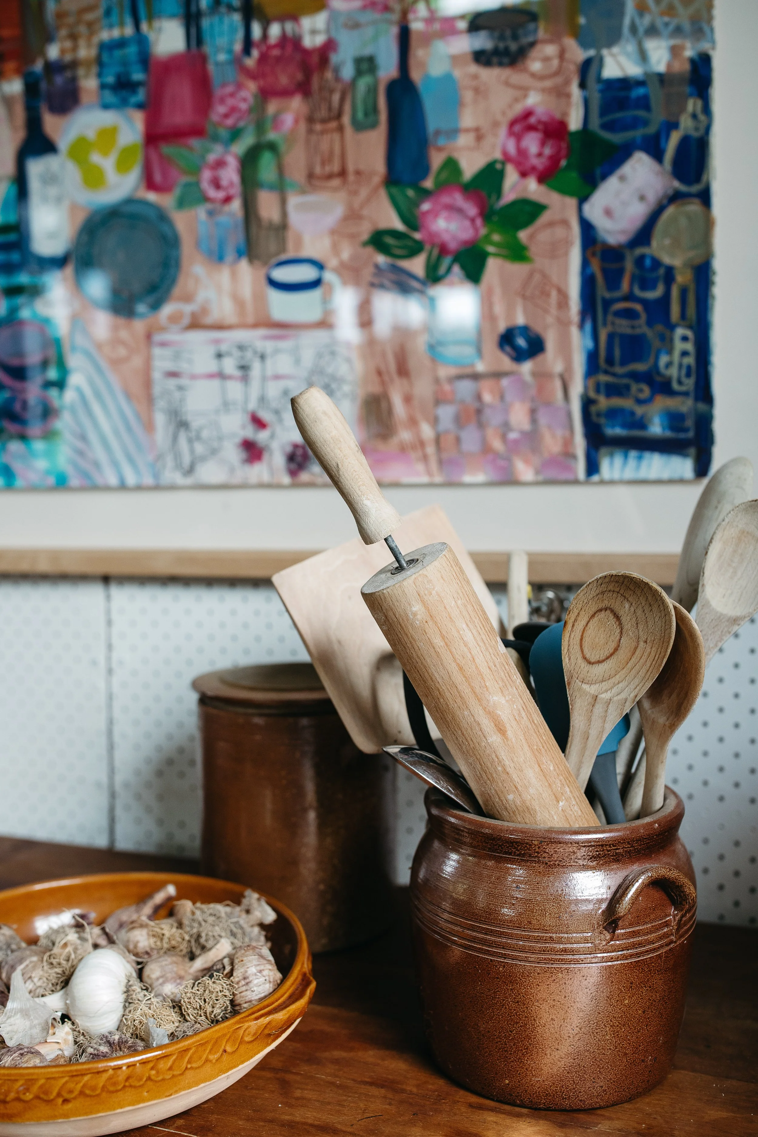A brown ceramic container holding wooden kitchen utensils, a rolling pin, and a spatula on a wooden table. In front, there is a bowl filled with garlic cloves and garlic bulbs. A painting of a colorful table setting hangs on the wall in the background.