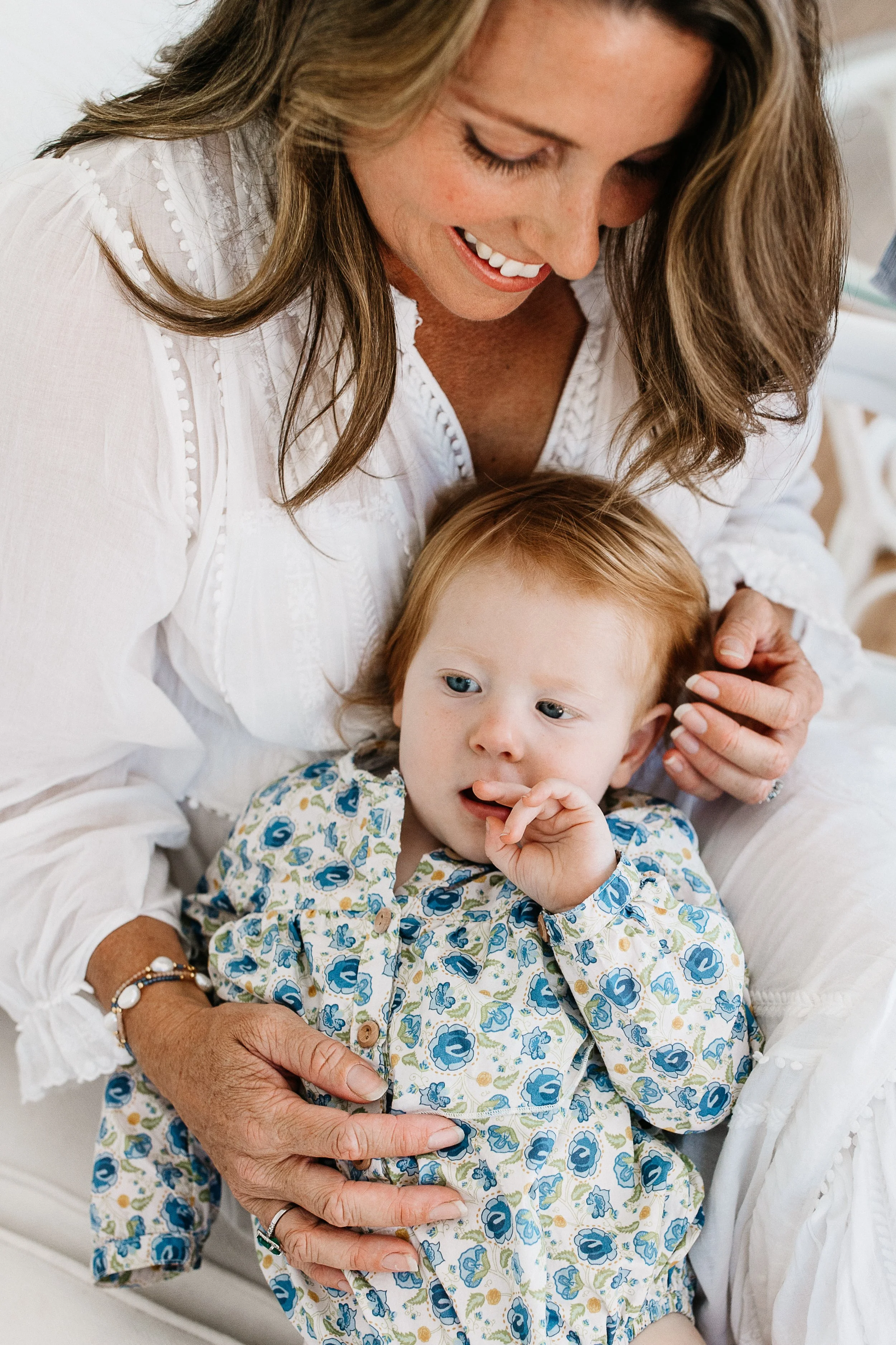 A woman with long brown hair and a white blouse smiling while holding a young child with red hair and blue eyes, dressed in a floral outfit, sitting on her lap.
