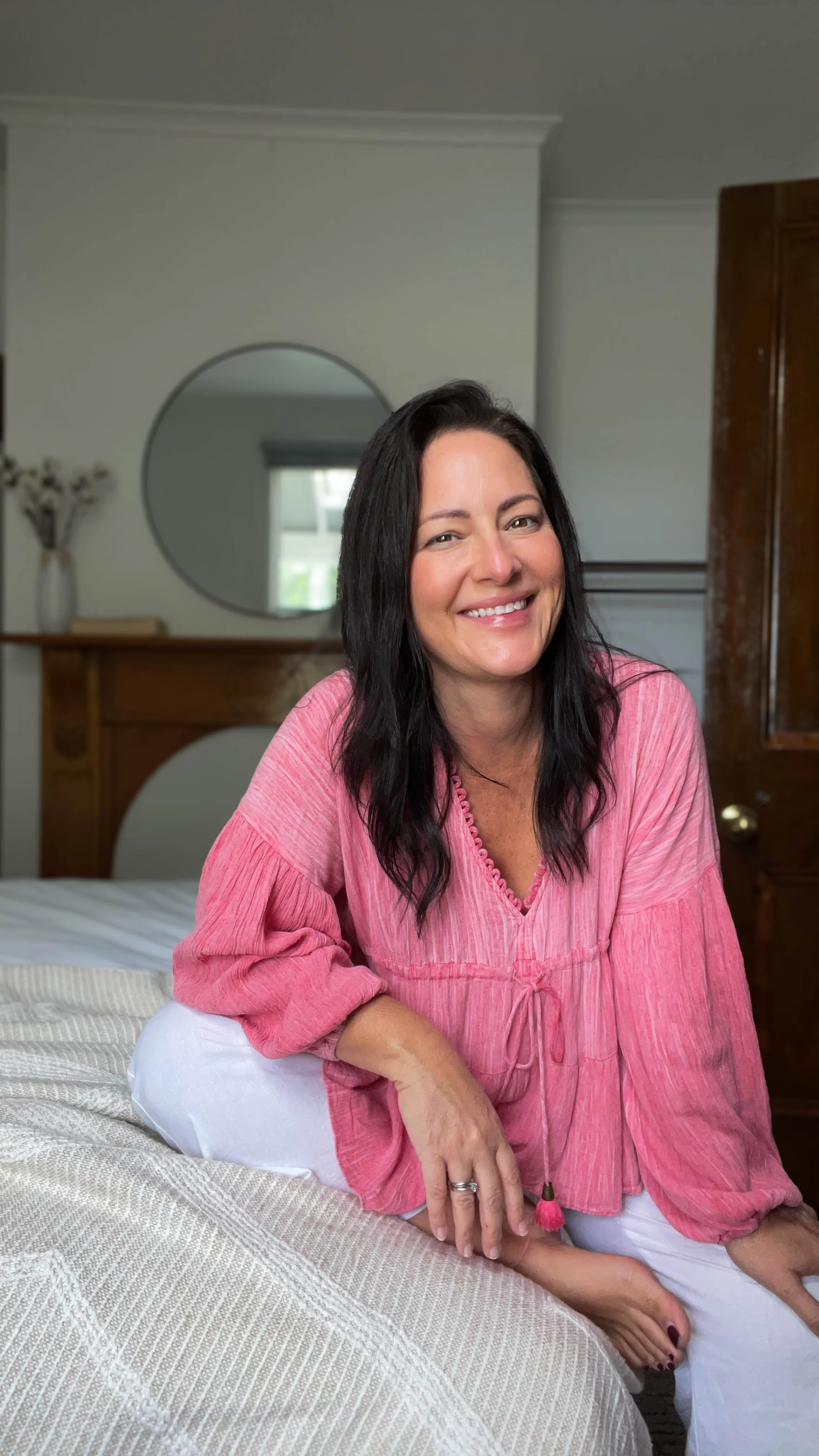 A woman with long dark hair, smiling, wearing a pink top and white pants, sitting on a bed in a bedroom with a mirror, vase, and wooden furniture in the background.