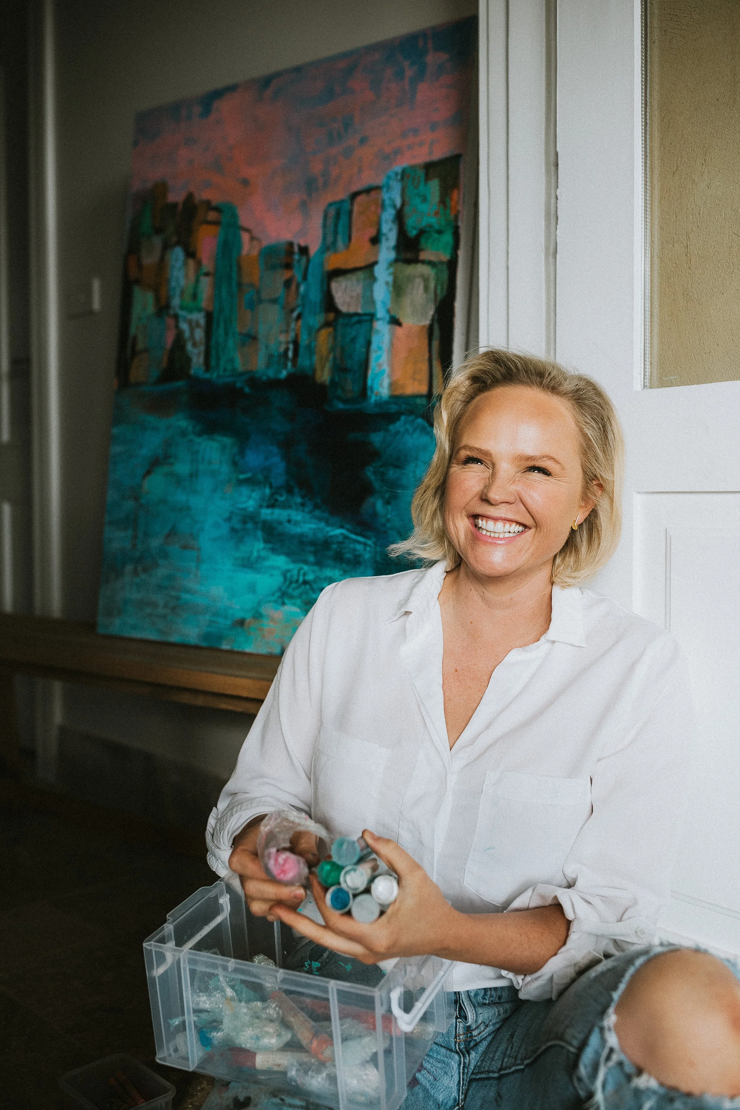 A woman with blonde hair and a white shirt smiling while sitting on the floor holding a container of paint tubes, with a large colorful abstract painting in the background.