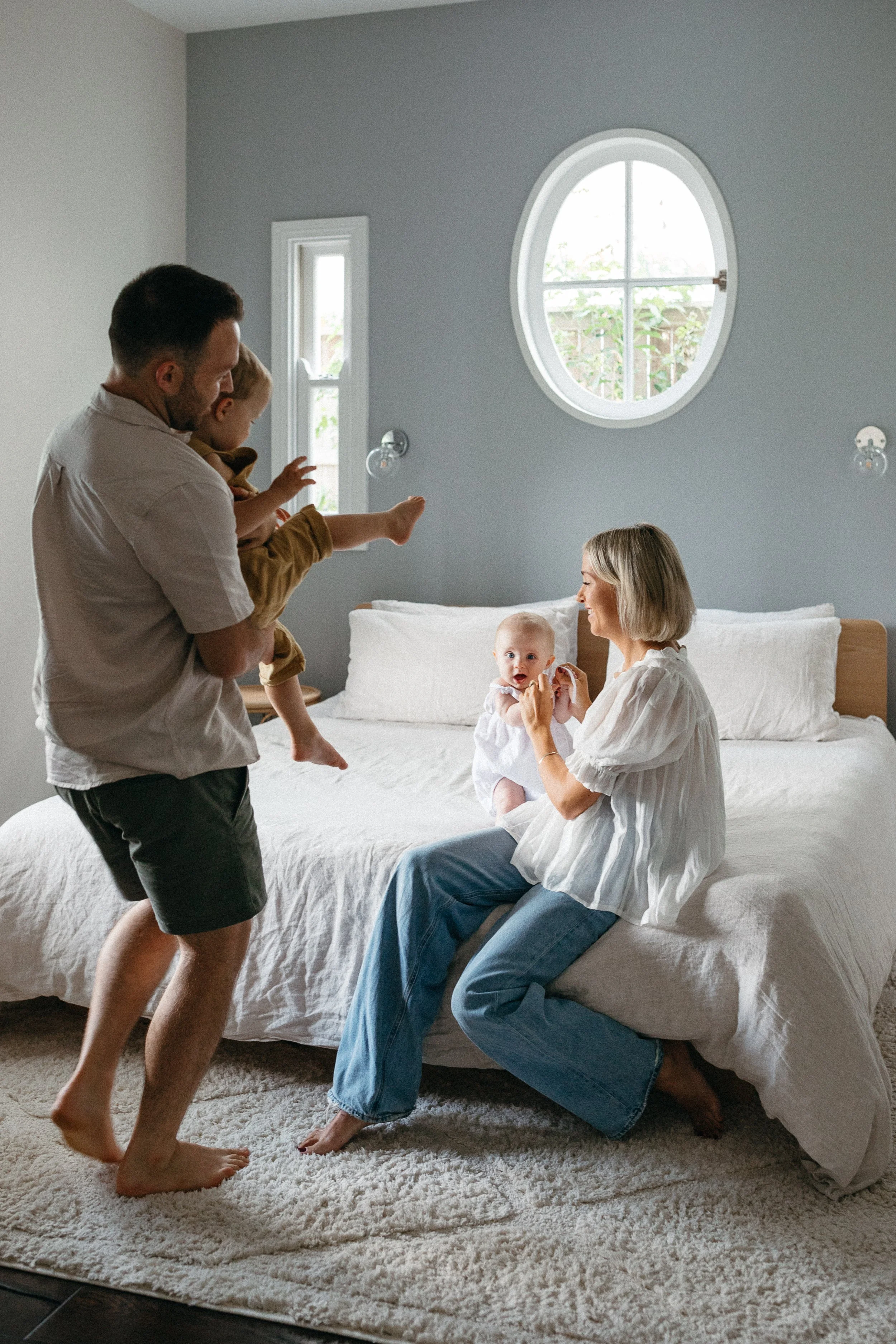 Family in bedroom playing with father holding a young child, and mother sitting on bed with another baby.