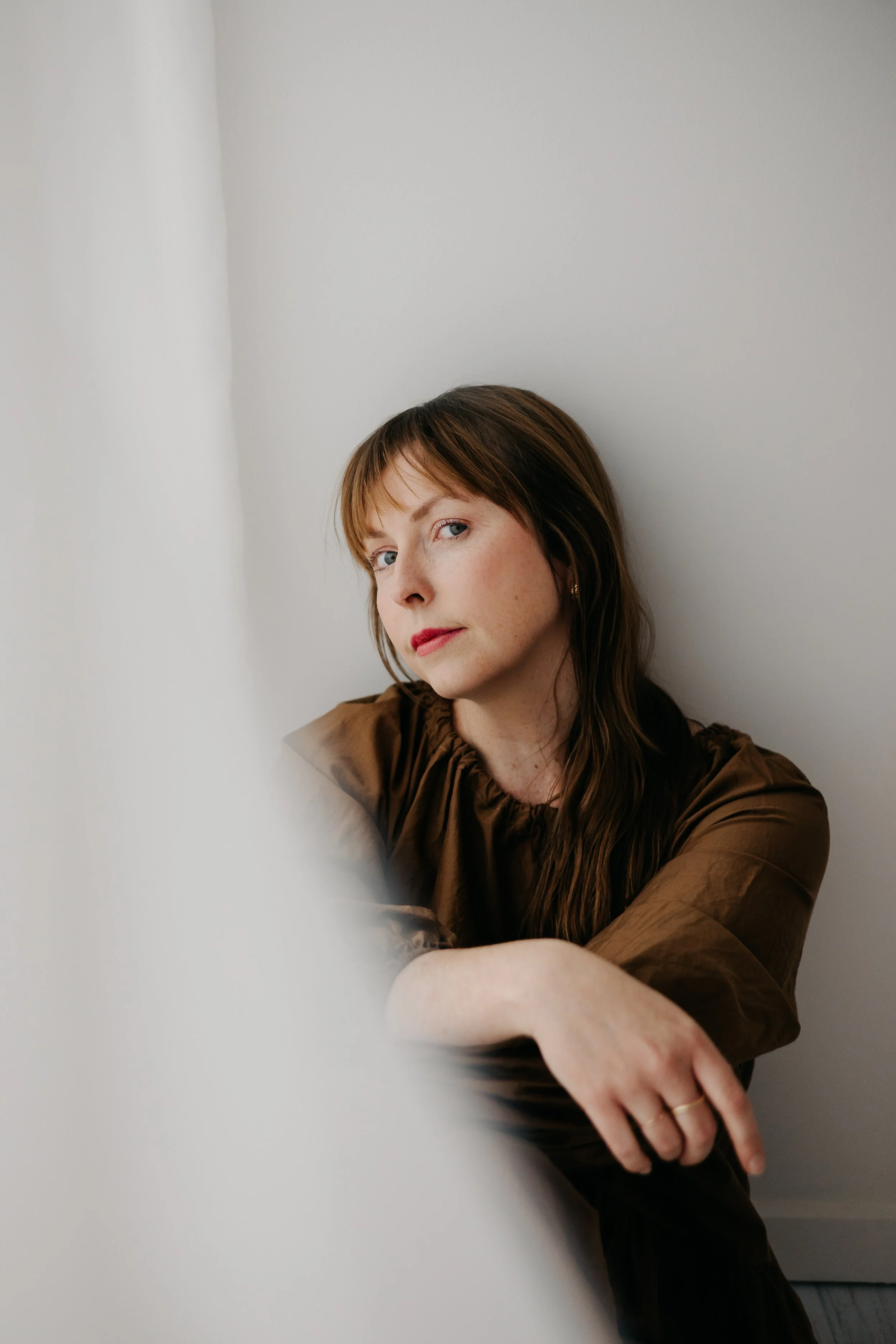 A woman with red hair and blue eyes sitting against a plain white wall, wearing a brown shirt and looking slightly to the left.